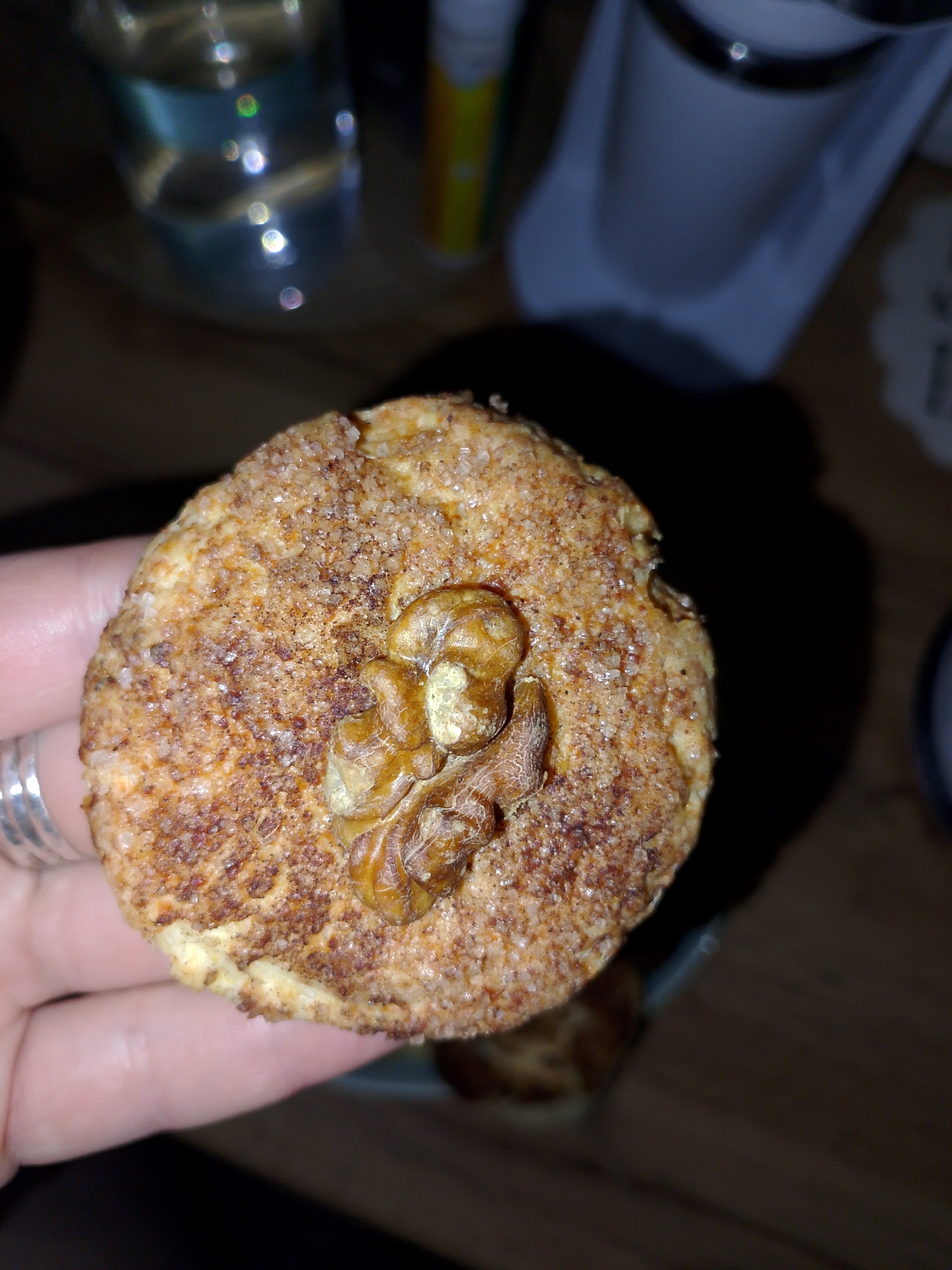 A hand holds a round, flat, baked good topped with granulated sugar and a walnut. The baked good is light brown, and the walnut is golden brown. A blurred drinking glass is in the background, as well as a white mug. The image is taken indoors, in a dimly lit area. The location in Izhevsk, Russia cannot be determined from the given image.