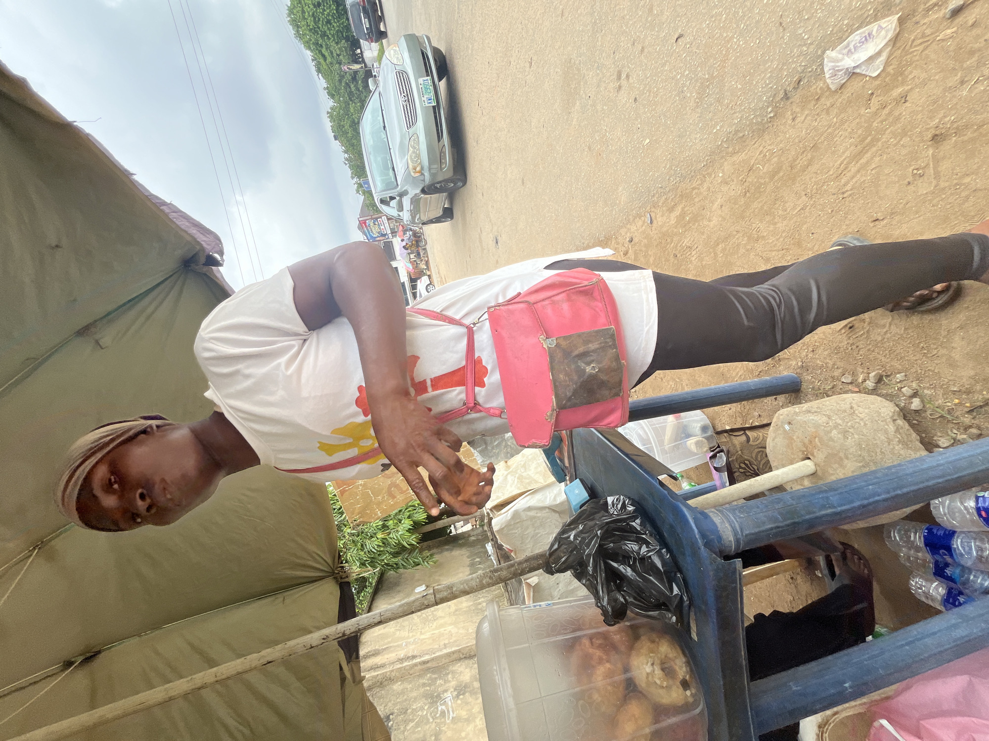 A young woman is standing under a khaki-colored umbrella next to a blue table on a dirt road in Abuja, Nigeria. She is wearing a white t-shirt, black leggings, and a beige headscarf. A pink purse with a metallic flap is slung over her shoulder. On the table, a clear plastic container holds what appear to be fried dough balls, and a black plastic bag is also present. Several bottles of water are on the ground beneath the table. In the background, a silver sedan is driving on the road, and trees and buildings are visible. The sky is overcast.