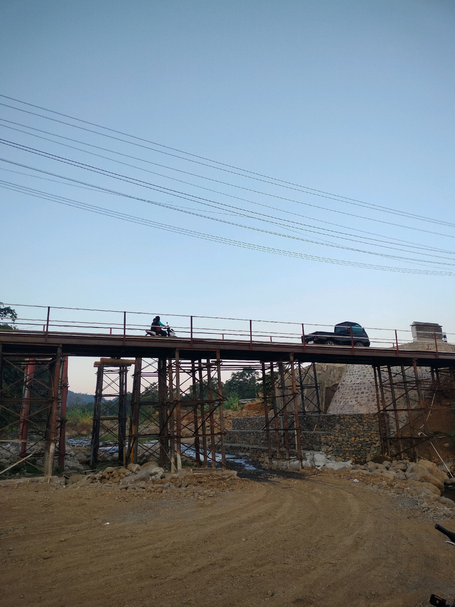 A wide shot captures an unfinished bridge over a riverbed in Tersono, Indonesia, under a clear, pale blue sky. Multiple dark power lines stretch across the upper portion of the frame. The bridge itself is constructed of rusty metal beams and supports, with a dark metal railing lining its edges. A person rides a scooter across the bridge from left to right, silhouetted against the sky. To the right of the scooter, a black pickup truck is also traveling across the bridge. The riverbed below is a mix of water, rocks, and construction debris. The ground in the foreground is a dirt track with scattered rocks and sand. The background shows dense foliage and hills.