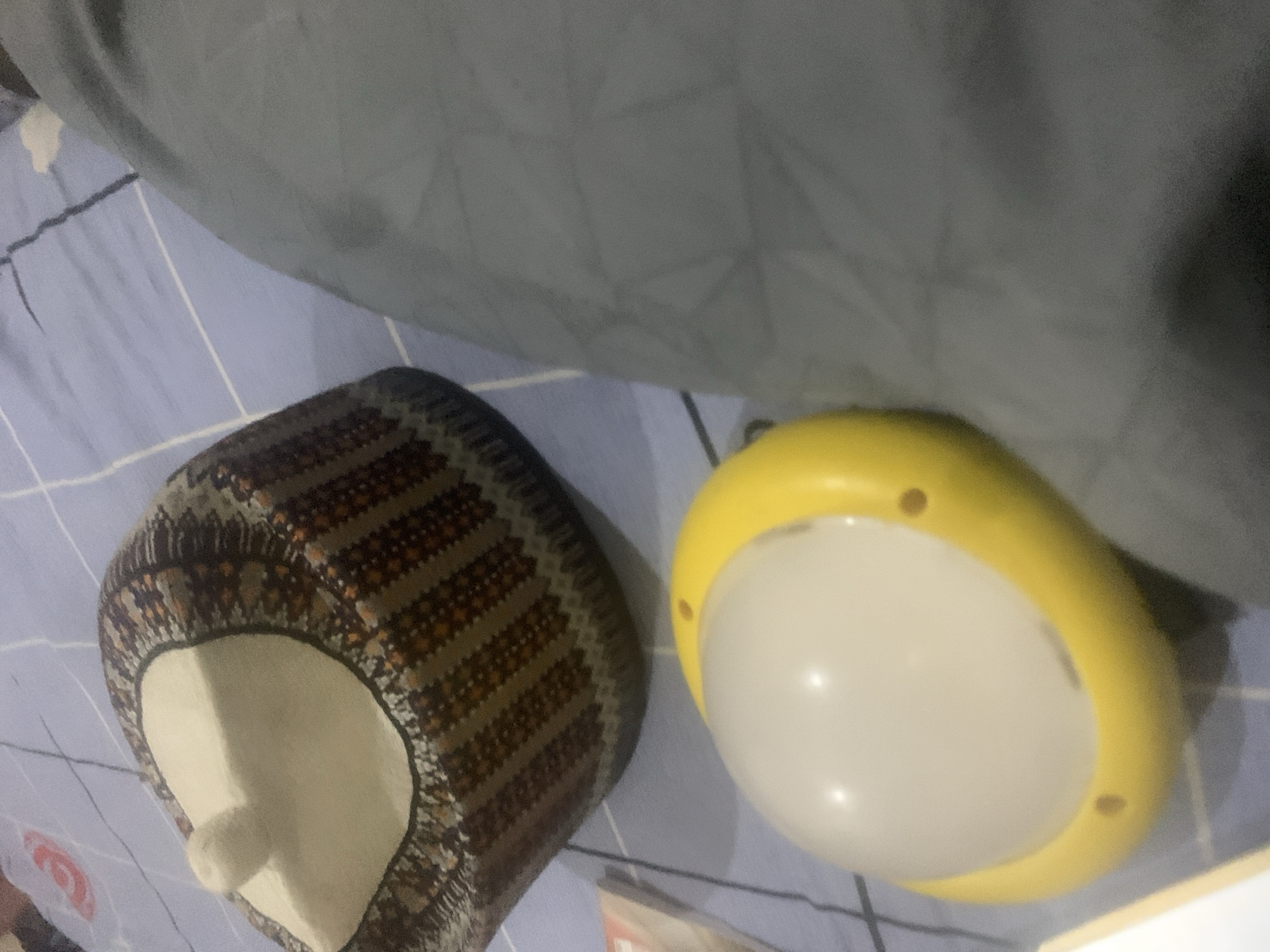 The image shows a top-down view of a patterned cap and a yellow and white portable lamp on a blue and white striped bedsheet. The cap has a white top with a small pom-pom and a patterned brim. The lamp is round with a yellow base and a white dome. A gray fabric is visible to the right of the objects. The lighting suggests an indoor setting. The location is identified as Jalingo, Nigeria, though no specific landmarks are visible in the image to confirm this.