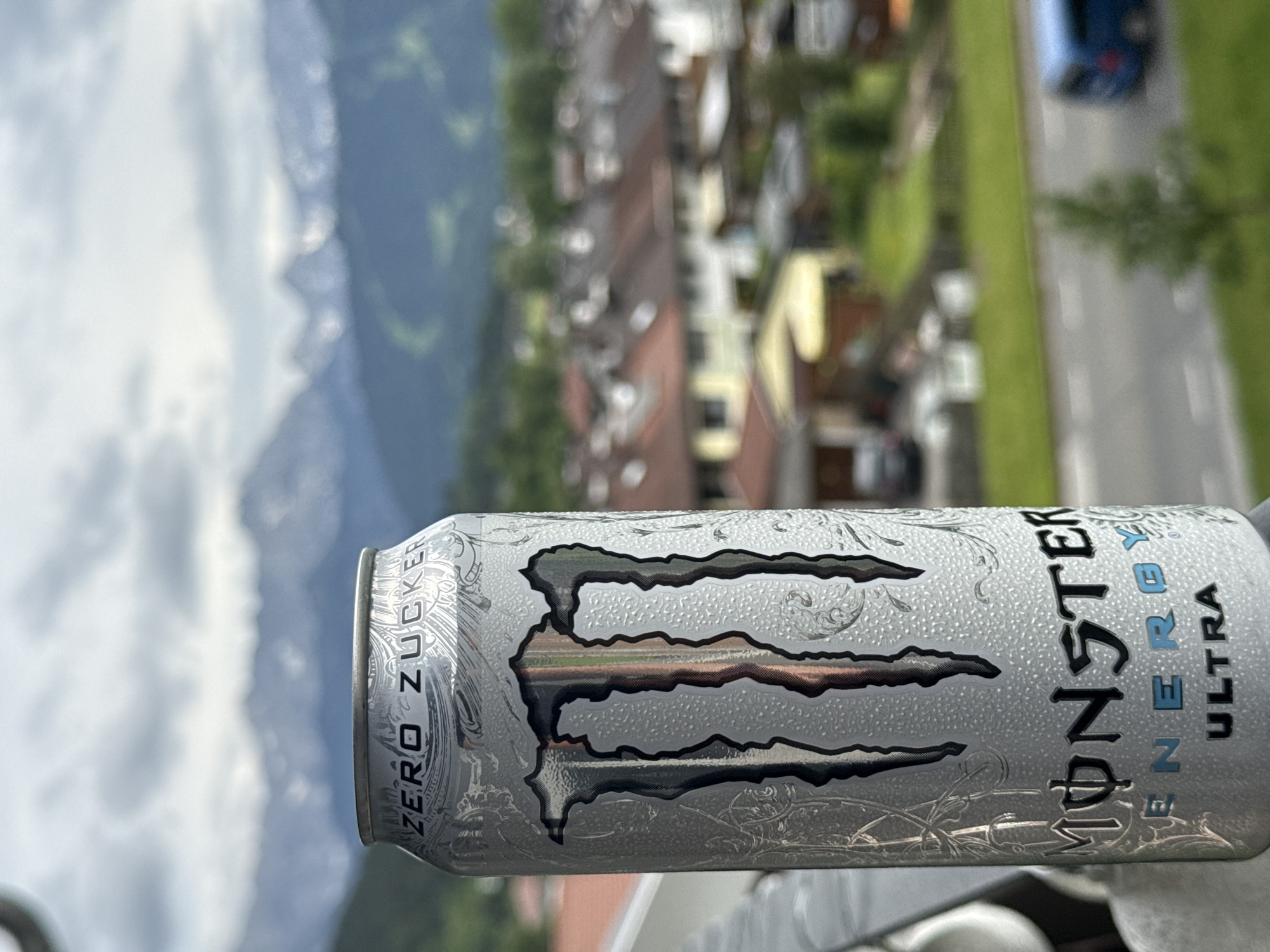 A Monster Energy Ultra can is in the foreground, positioned at an upward angle. The can is silver with the iconic Monster logo, "MONSTER ENERGY," and "ULTRA" visible. The text "ZERO ZUCKER" is also present on the can. The background is blurred, showing a residential area with houses with reddish-brown roofs. Beyond the houses, mountains are visible under a cloudy sky. The scene is set in Garmisch-Partenkirchen, Germany.