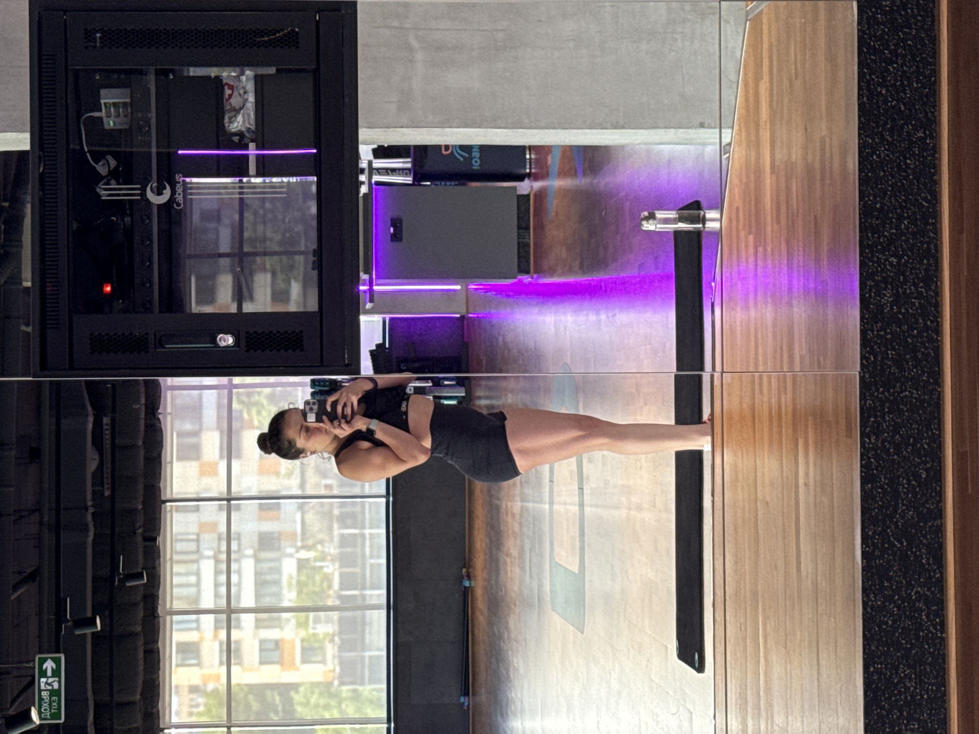 A woman is reflected in a gym mirror with exercise equipment, wooden floors, and a black tiled pillar. Outside the window are buildings. It is not possible to verify from this photo that the location is in Moscow, Russia.