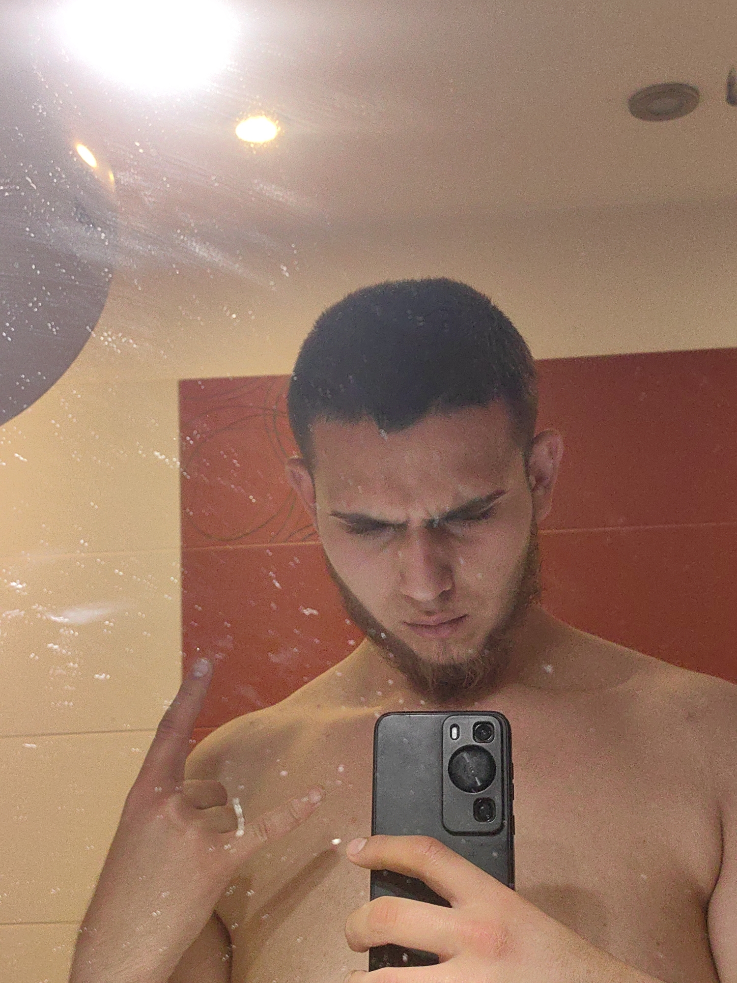 A fair-skinned man is taking a selfie in a bathroom mirror. He has dark hair and a short beard. He's holding a black smartphone in his right hand and making the "rock on" hand gesture with his left. He appears to be frowning slightly. The mirror is speckled with water droplets. Behind him, the bathroom wall has a two-tone color scheme, with a reddish-orange panel and a beige panel. The ceiling has recessed lighting. There is no clear information about the city of Sofia or Bulgaria from this image.
