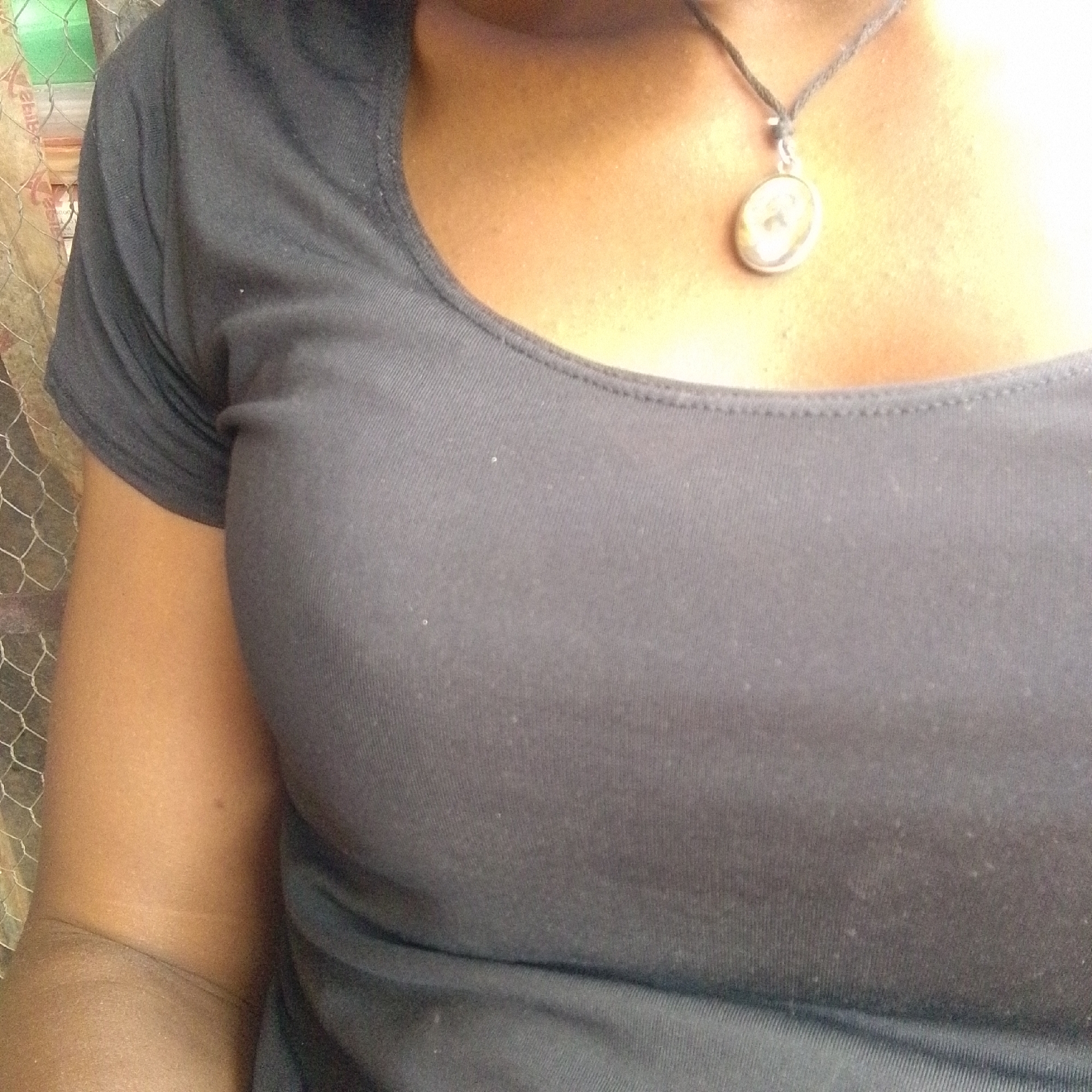 A close-up shot shows the upper torso of a dark-skinned person wearing a dark, short-sleeved shirt. The person is also wearing a necklace with a round pendant. A chain-link fence is visible on the left side of the image. Information about the location or any specific context within the city of Jos, Nigeria, is not discernible from the image.