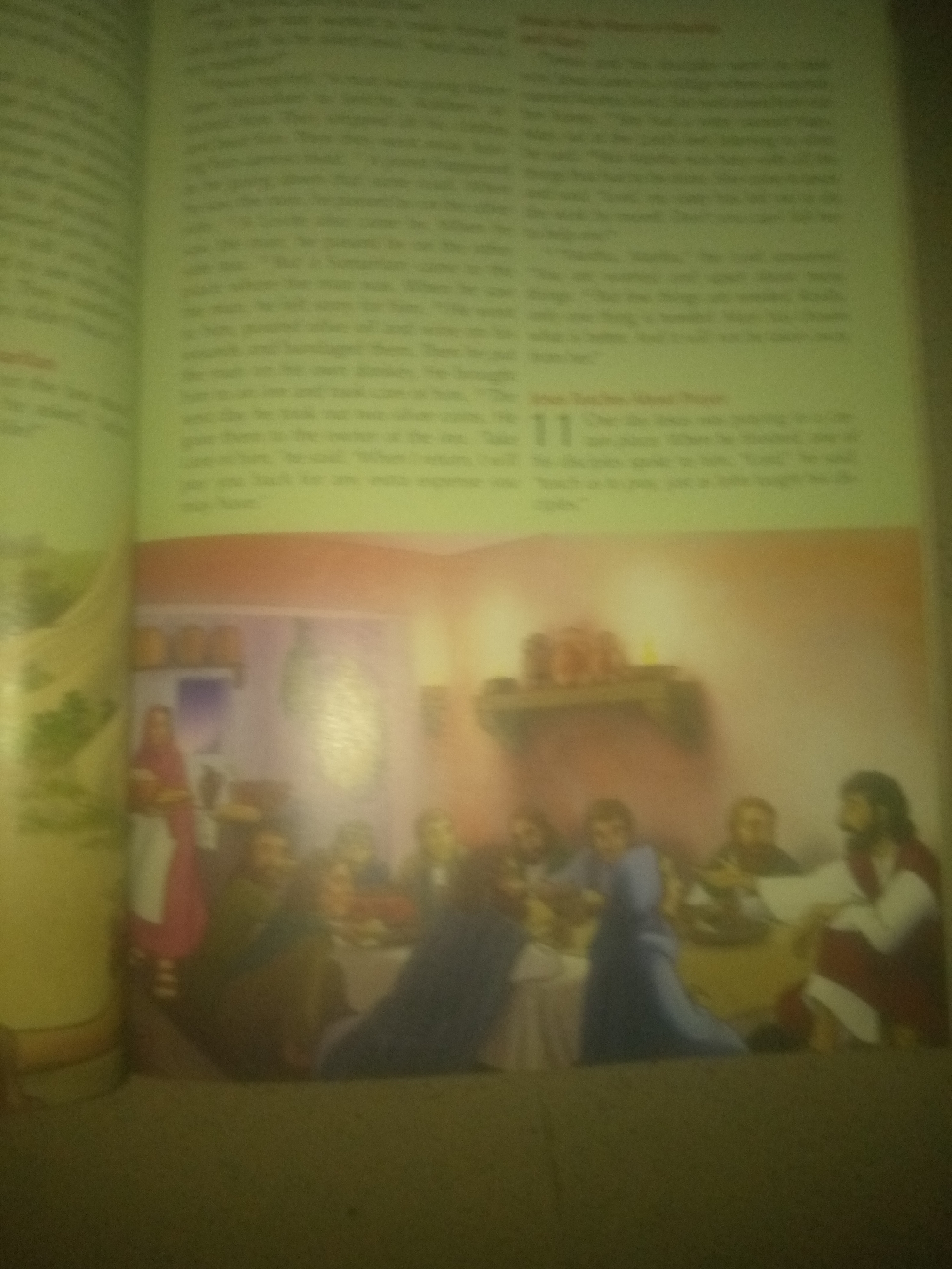 The image is a photograph of an open book. The left page displays text, and the right page contains text and an illustration. The illustration depicts a biblical scene with several figures gathered around a table, possibly for a meal. In the foreground of the illustration, there are multiple individuals seated, while in the background, a woman in a red garment stands near a shelf with some items. The overall scene appears to be indoors, possibly in a dining or gathering area. The text on both pages is blurry and illegible, as is the image due to the low resolution and focus of the photograph. There is no visible connection to Jauro Yinu, Nigeria.