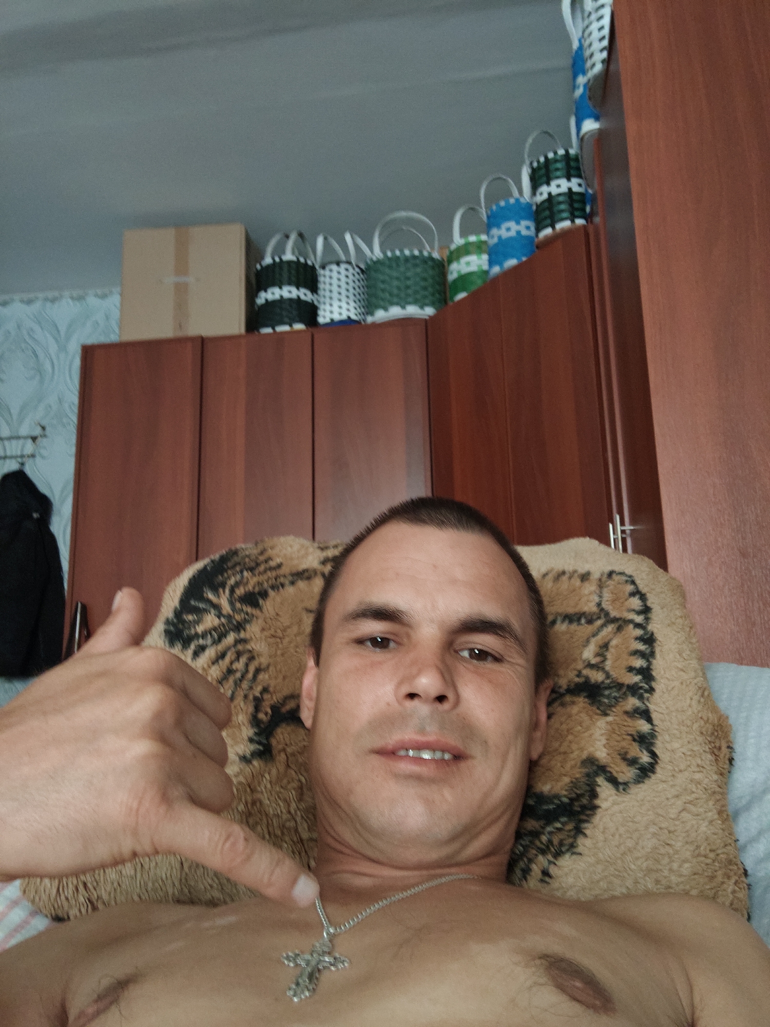A shirtless man with short brown hair and a cross necklace is lying on a tan, patterned pillow. He is looking at the camera and has his left hand raised in a gesture. In the background, there are wooden cabinets and several woven baskets on top of them. A brown cardboard box is also visible on top of the cabinets. A black jacket hangs on a rack to the left of the cabinets. The scene appears to be indoors, likely in a residential setting. The image was taken in Курашим, Russia.