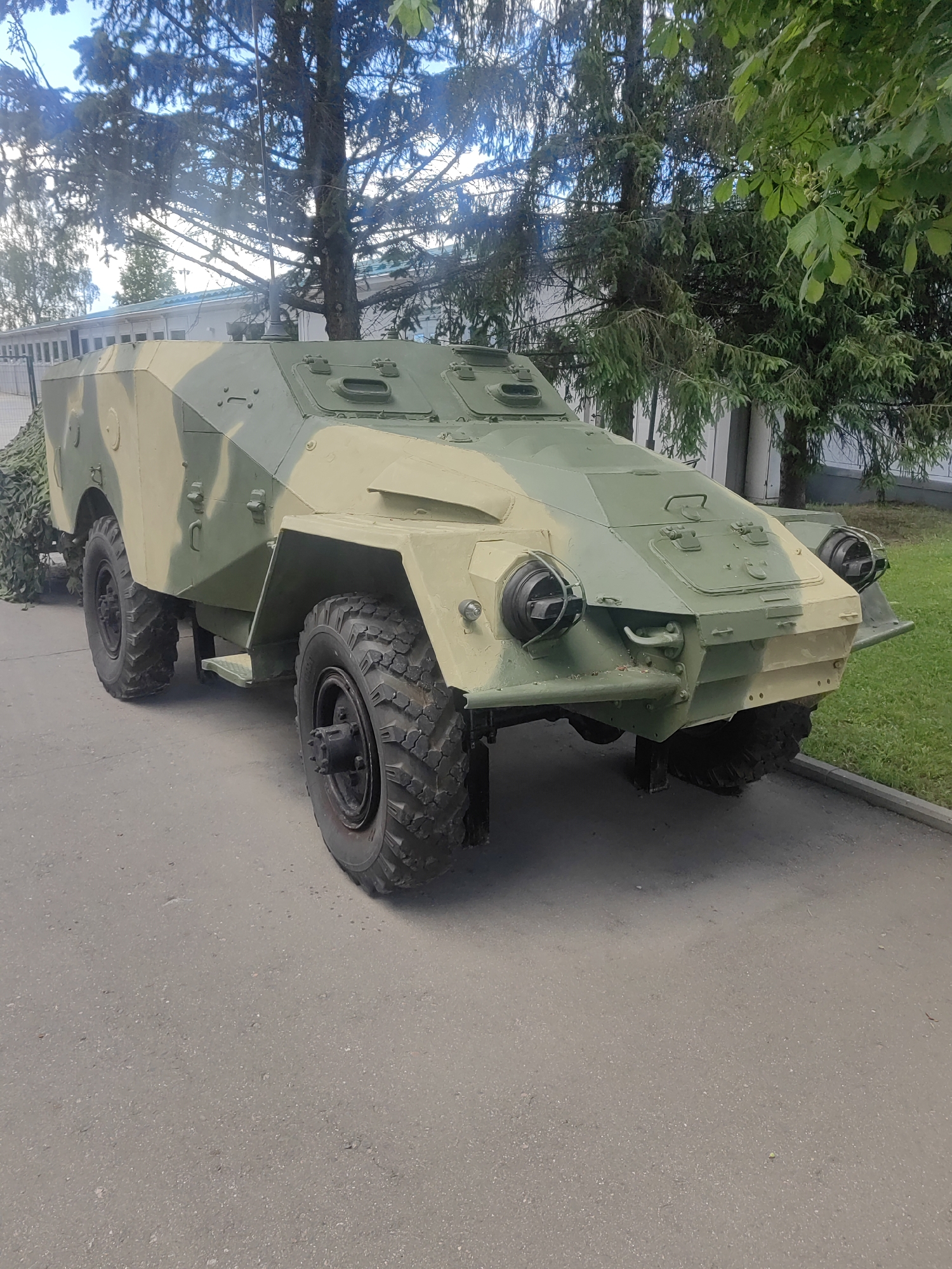 The media file displays an outdoor medium shot of a camouflaged military vehicle, likely an armored car, parked on an asphalt surface. The vehicle is painted in a disruptive camouflage pattern of light green and tan. Its design is angular and rugged, featuring large, knobby tires, prominent headlights with protective cages, and visible hatches on its roof. A metal step is attached to the side of the vehicle. Behind the vehicle, a building with a white facade and windows can be seen, partially obscured by trees. The trees are lush with green foliage, suggesting a warm-weather setting. The sky is visible in the upper portion of the image, appearing partly cloudy. The lighting suggests it is daytime, possibly late morning or afternoon. The vehicle appears stationary, and no people are visible. The overall scene is one of a static display, potentially at a museum or exhibition, given the military equipment and the presence of a building that could be part of such a facility. The location context suggests this could be in Kubinka, Russia. No visible text is present in the image.
