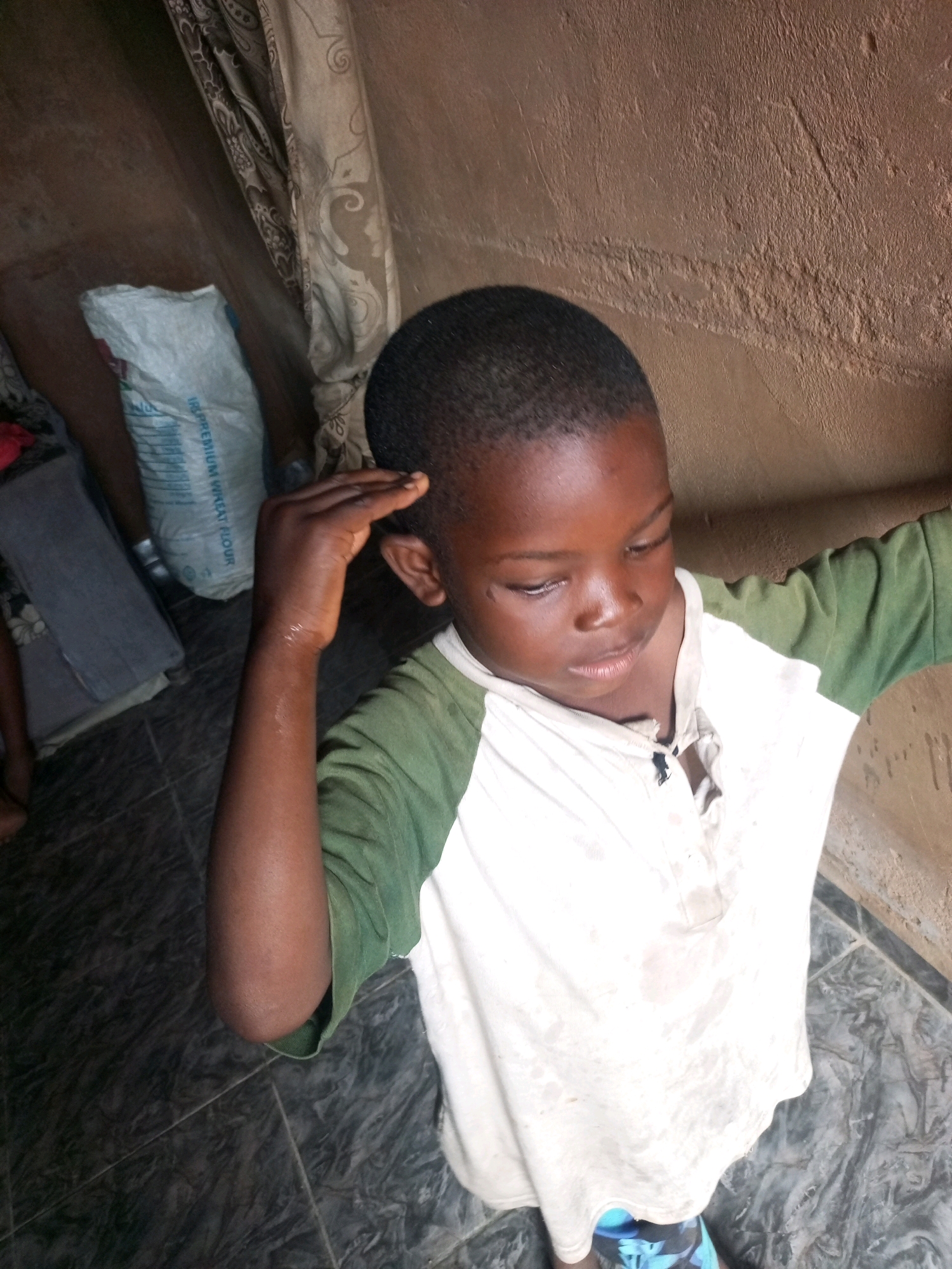 In Tempure, Nigeria, an overhead shot captures a young boy standing inside what appears to be a modest dwelling. The boy, with dark skin and close-cropped hair, is dressed in a white t-shirt with green sleeves, which appears stained. He is looking downwards, with his right hand touching his forehead. To the left of the boy, a large white bag, possibly containing flour, is visible leaning against a textured, brownish wall. Further to the left, a gray fabric or mat is folded. The floor is tiled with a dark, mottled pattern. The overall environment suggests a simple, possibly rural or less developed urban setting.