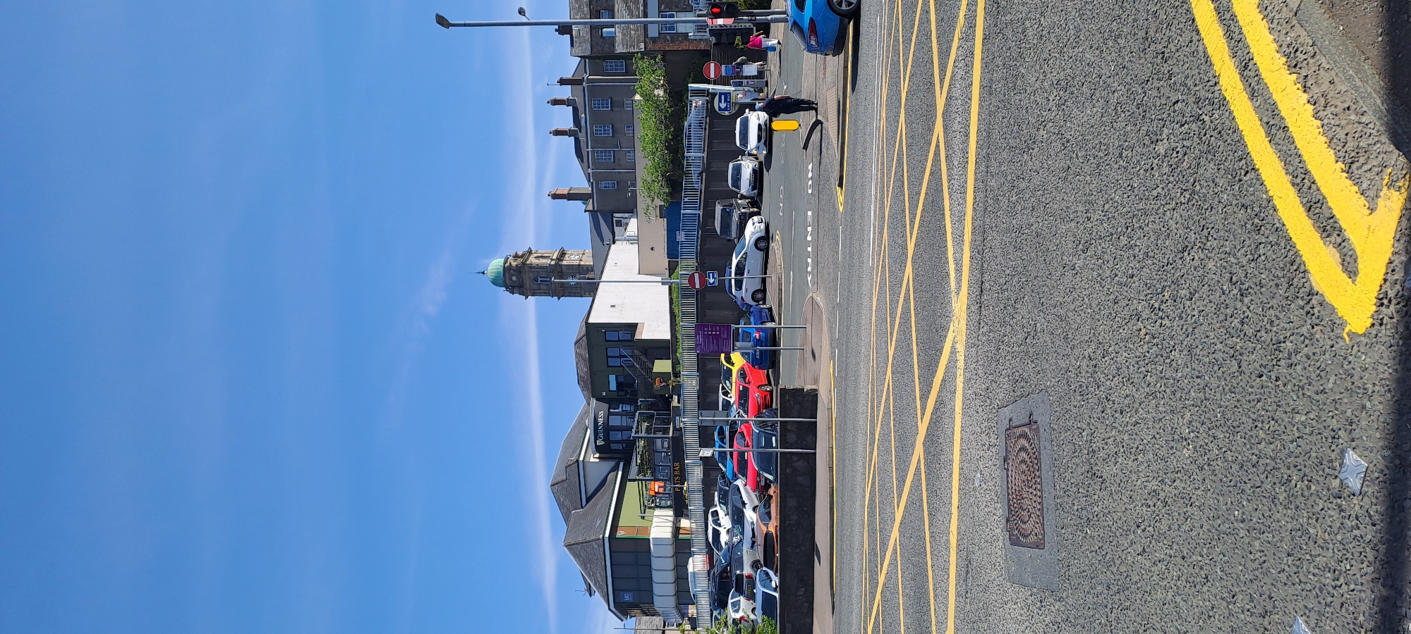 In Enniskillen, United Kingdom, a scene unfolds on a clear, sunny day, featuring a street intersection with prominent yellow box junction markings. In the foreground, the asphalt is textured with a distinct pattern, interrupted by a metal manhole cover and several reflective road studs. A lone person, dressed in dark clothing, stands near a yellow barrier in the middle ground, appearing to be in motion.
Beyond the intersection, a parking lot is filled with various vehicles, including white, blue, red, and yellow cars, as well as a dark-colored SUV. A low stone wall separates the parking lot from a raised area where a building complex stands. This complex features a multi-story structure with a dark roof, a green facade, and a prominent "GUINNESS" sign. To the right of this building, a tall, square tower with a green, domed roof and visible clock faces rises against the bright blue sky, suggesting a landmark or public building. Further to the right, residential-style buildings with brick chimneys are visible.
On the street, traffic signals are present, with one showing a red light. Other signage includes "ONE WAY" arrows and a red circular sign with a diagonal line, indicating "NO ENTRY." A blue car is parked on the right side of the street. The overall environment suggests an urban setting with commercial and residential elements, under a cloudless blue sky.