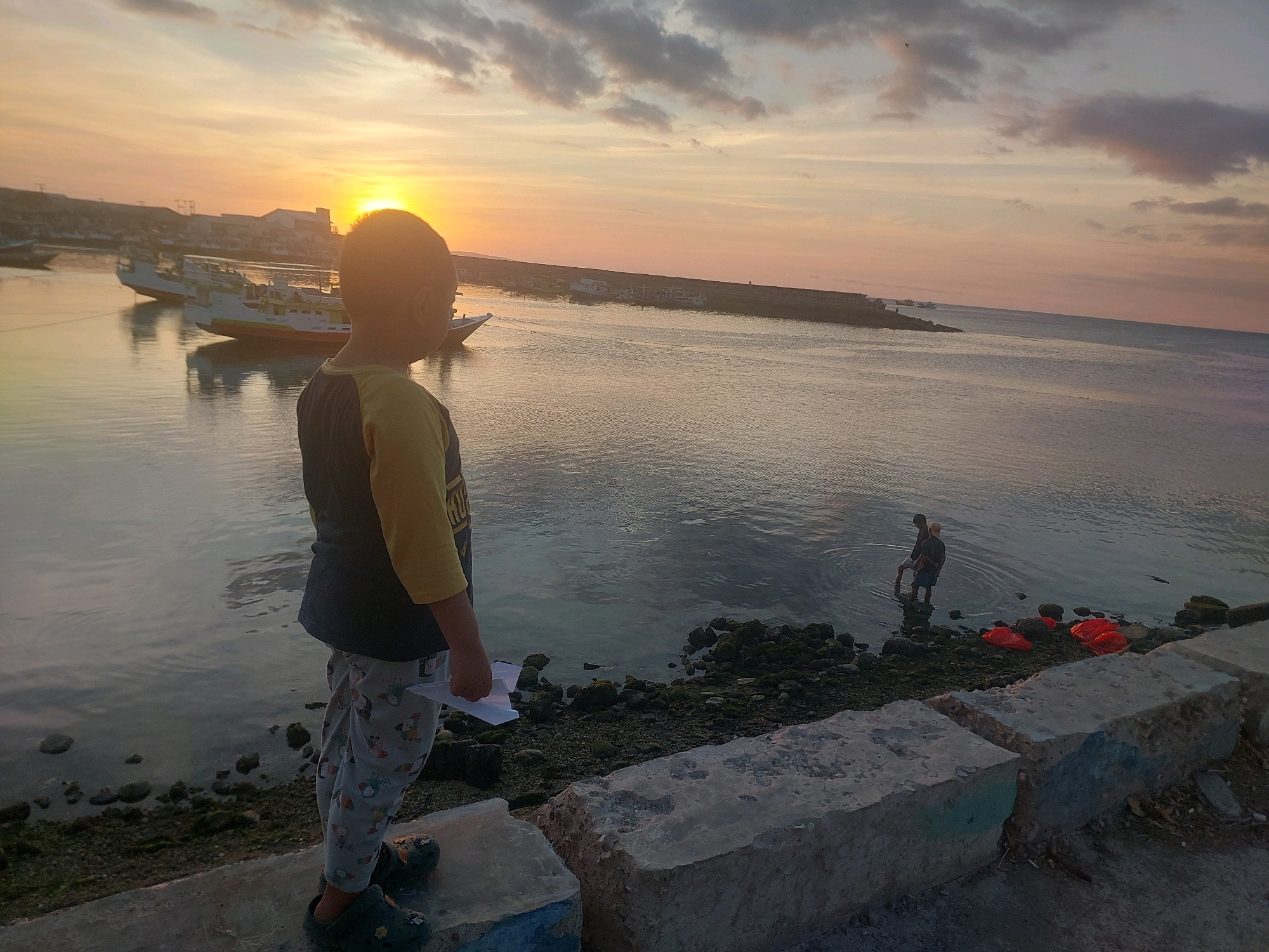 A young child stands on a concrete embankment, looking out at a calm body of water during sunset. The child, wearing a yellow and dark t-shirt, patterned pants, and dark clogs, holds a piece of white paper. In the distance, several boats are docked, and a man is wading in the shallow water near the shore. The sky is filled with soft clouds, painted with the warm hues of the setting sun. The scene appears to be in a coastal area, possibly a harbor or bay, in Kupang, Indonesia, given the characteristic landscape and the presence of traditional boats. The overall atmosphere is peaceful and serene, capturing a moment of quiet observation against a beautiful natural backdrop.