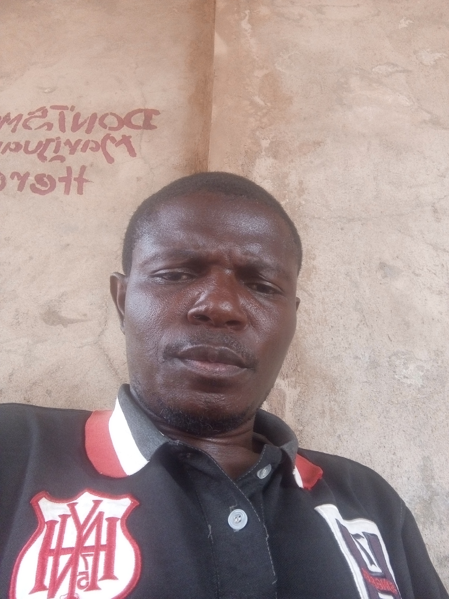 A medium shot depicts a Black male with short, dark hair and a mustache and goatee. He appears to be inside a building with a rough, beige wall, possibly made of concrete, in the background. The wall has red lettering, but it is largely obscured. The man is wearing a dark polo shirt with a white and red collar. There is a white and red emblem on the left side of his chest. The location appears to be within the city of Unknown, Unknown.