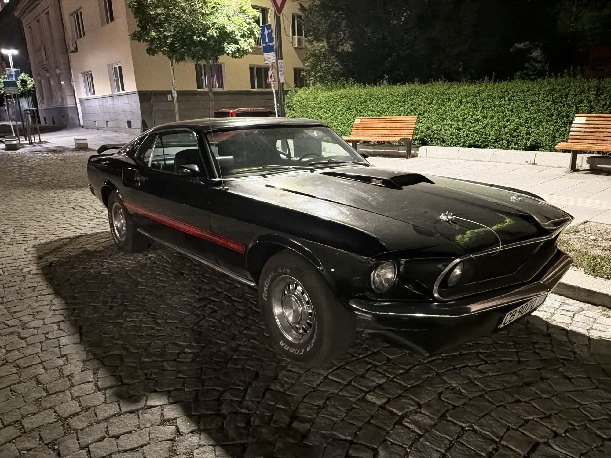 A black 1969 or 1970 Ford Mustang Mach 1 is parked on a cobblestone street at night in Sofia, Bulgaria. The classic car features a prominent hood scoop with "351" stenciled on it, chrome accents, and a red horizontal stripe along its side, where "Mach 1" is visible. Chrome hood pins secure the front of the hood. The tires display "COBRA RADIAL G/T" on their sidewalls. A Bulgarian license plate, "CB 9070 C", is mounted on the front bumper. The car is angled towards the right side of the frame, with its front facing slightly left.
The cobblestone street is unevenly illuminated by an unseen light source, casting long, distinct shadows from the vehicle and nearby objects. In the background on the left, a multi-story building with a light-colored facade and a grey ground floor is visible, featuring several rectangular windows. A lush green tree with light-colored bark stands between the building and the street. Further to the right, behind the car, a dense, well-maintained green hedge runs along a raised sidewalk. Two slatted wooden park benches are positioned along this hedge. Streetlights provide illumination, creating a contrast between bright and shadowed areas. A pole with a blue directional sign featuring an upward-pointing arrow and other smaller signs is visible on the left side of the street. The scene is devoid of people.