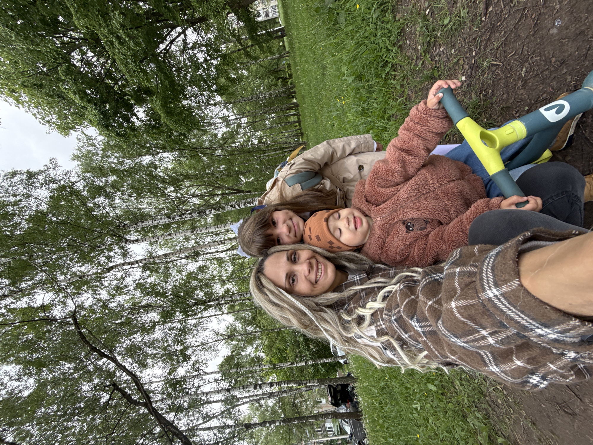 The image depicts a woman with blonde hair wearing a brown plaid shirt lying on the grass with two children, presumably her own, in what appears to be a park or grassy area. One child is wearing a beige jacket, while the other is wearing a brown, teddy bear-like jacket and an orange hat. They are lying close together, seemingly engaged in taking a selfie or group photo. A green and yellow object, possibly a child's toy or scooter, is also visible. The background features trees and vegetation, and there are buildings in the distance.
While the city of Rusanovo, Russia is mentioned, the location is not explicitly confirmed as such. However, assuming the location is accurate, the scene would then be a casual moment of leisure in a park environment within Rusanovo, involving a family engaging in a recreational activity.