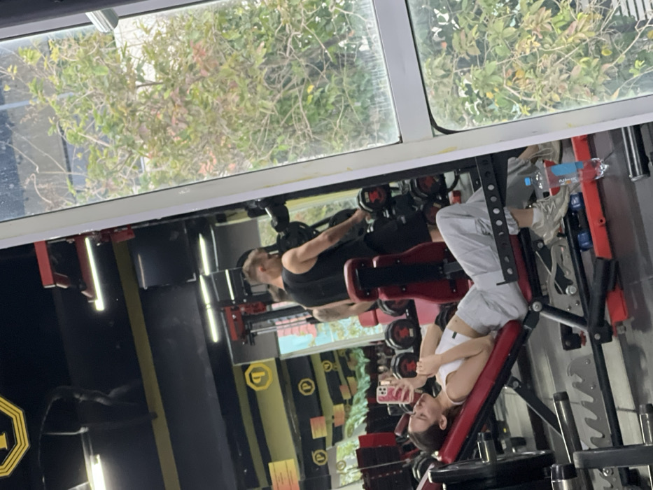 The image depicts an indoor scene within a gym, identified by 'P' logos and branding consistent with "Prime Fitness & Yoga," a facility found in Nha Trang, Vietnam. A young adult female is positioned in the foreground, lying reclined on a red and black adjustable weight bench. She wears a white sports bra, light grey sweatpants, and light-colored athletic shoes. Her right hand holds a pink-cased smartphone, which she appears to be viewing or using to take a photograph. A plastic water bottle and a small blue dumbbell are visible on the floor near her feet. In the background, reflected in a large mirror, a young adult male is seen standing near a rack of dumbbells. He wears a black tank top and black shorts and is interacting with the dumbbells. Another male figure with a tattooed arm is partially visible behind him, also in the reflection. The right side of the image features a large window with white framing, offering an outdoor view of green trees and foliage under natural light. The gym interior features black walls with horizontal yellow stripes and yellow 'P' logos.