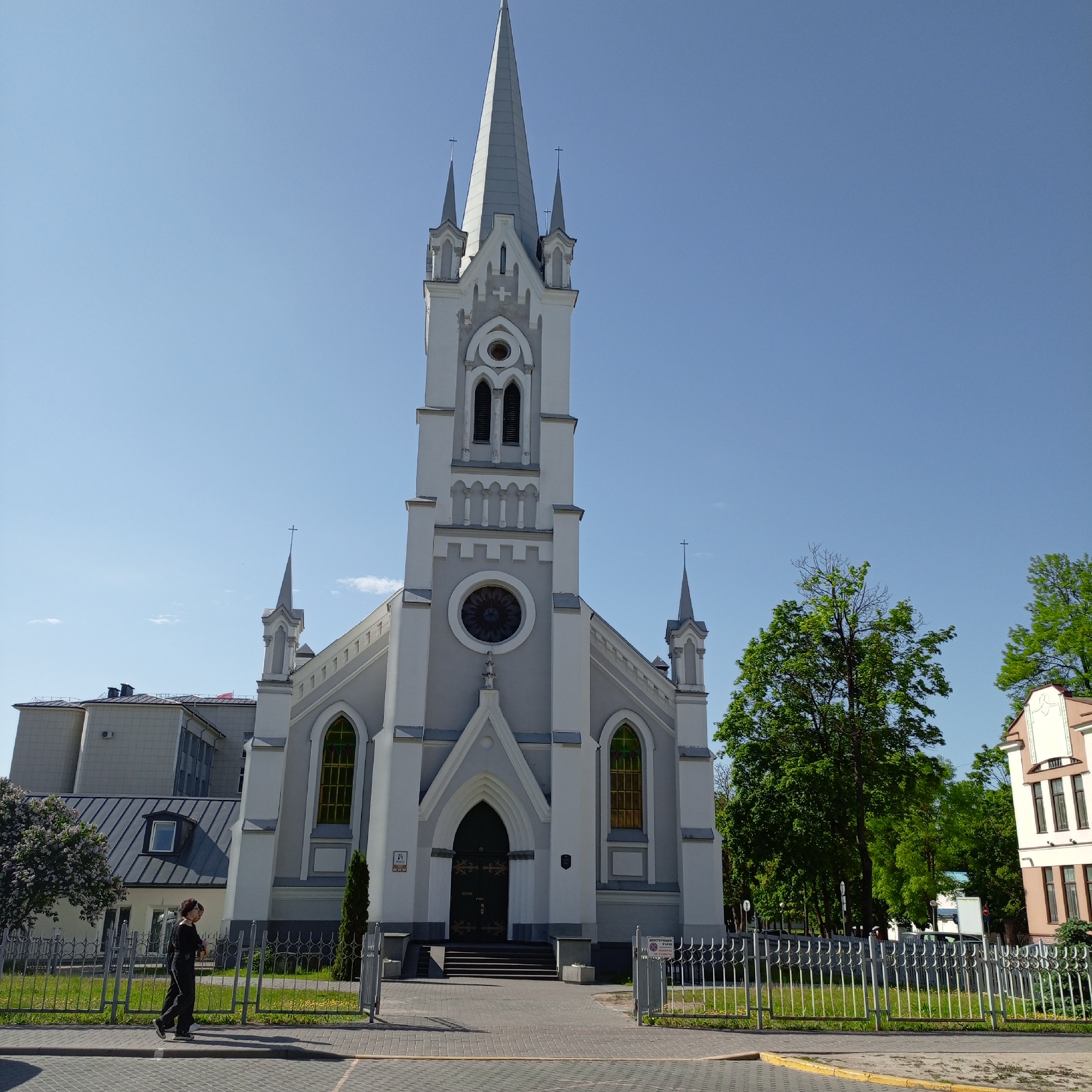 The image shows a large, gray and white church with a tall steeple, located in an urban environment. The church has several arched windows and a prominent rose window above the main entrance. The facade is detailed with Gothic-inspired architectural elements. A metal fence surrounds the church grounds. To the left of the church is a modern building, and to the right are trees and another building. A person is walking on the sidewalk in the lower left corner of the frame. The sky is clear and blue.
