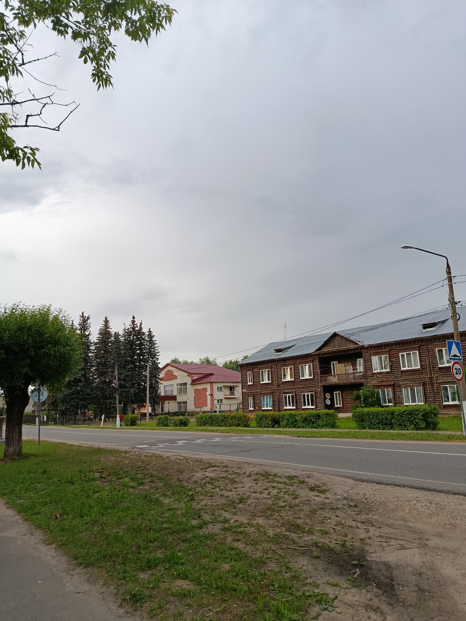 The image shows a street scene in the city of Vereya, Russia, on an overcast day. In the foreground, a paved sidewalk runs alongside a grassy area and a road. On the other side of the road, there is a row of buildings. The closest building is a large, two-story wooden structure with a dark, pitched roof and many windows. To the left of the wooden building is a smaller, lighter-colored building with a red roof. Behind the buildings, a line of tall evergreen trees can be seen. A street lamp and several road signs are visible near the wooden building, including a speed limit sign indicating 20. The sky is a uniform grey, suggesting cloudy weather.
