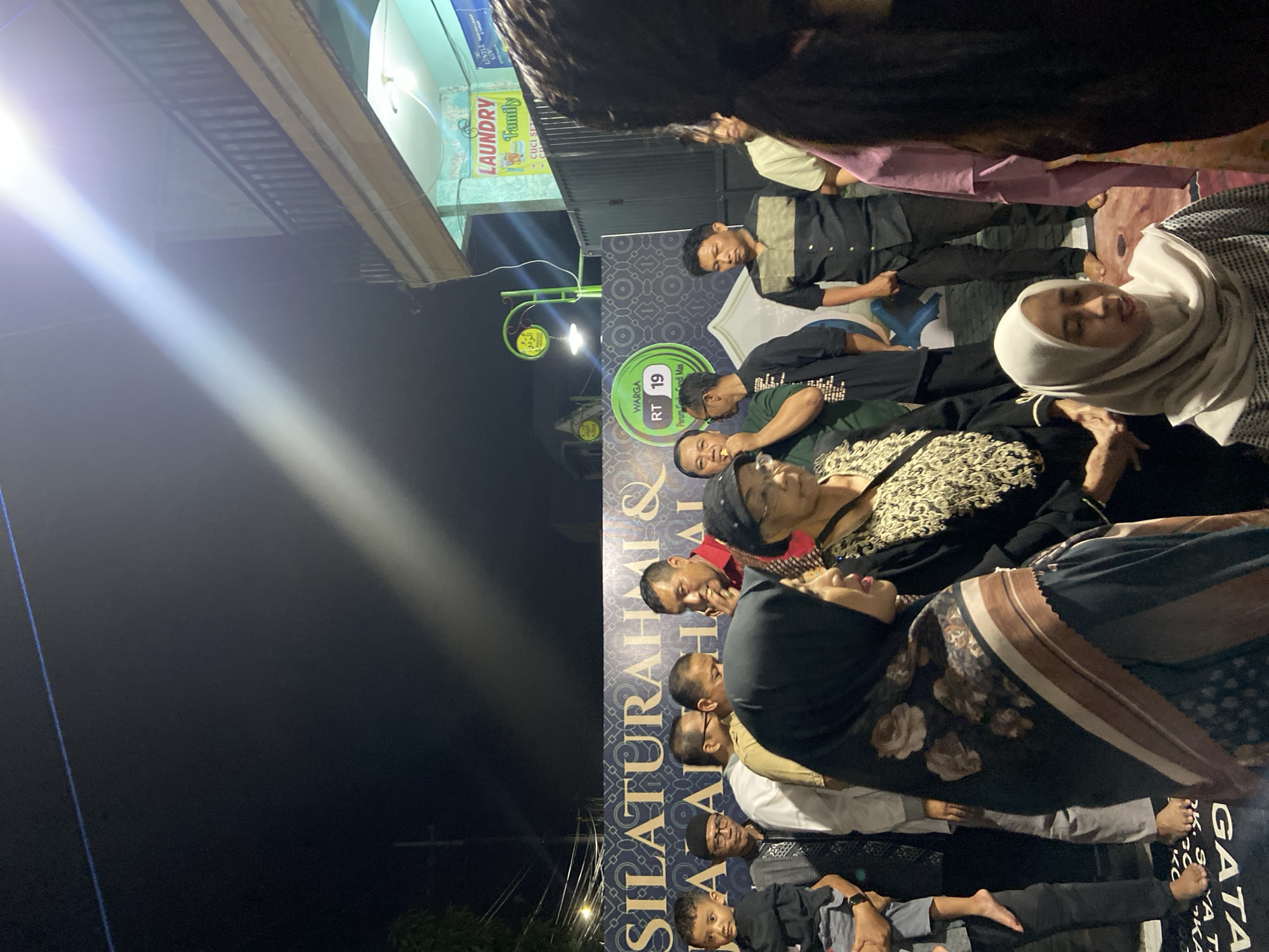 The image shows a group of people gathered outdoors at night in Gelam, Indonesia. They are in front of a banner that reads "SILATURAHMI & HALAL BI HALAL WARGA RT 19 Perum Gandi Mas K. SYA T GATA." Individuals, including women in hijabs, are seated or standing closely together. Above them is a sign for a "LAUNDRY Family" business. The scene appears to be a community gathering or celebration.