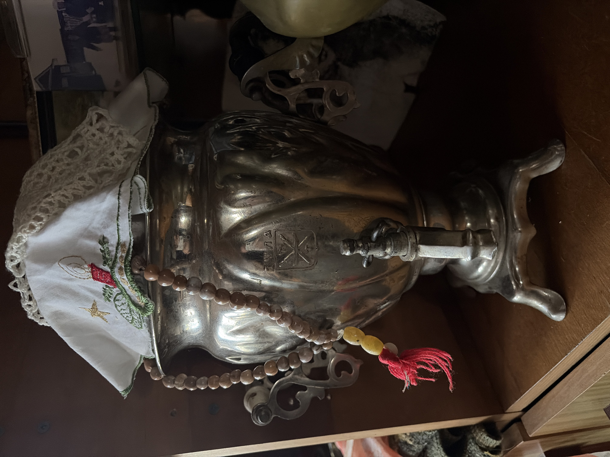 An antique, silver-plated samovar sits on a wooden shelf. It is decorated with beads and a red tassel. A white cloth with embroidered Christmas decorations is draped over the top. The samovar is placed in the city of Mamadysh-Akilovo, Russia, on a wooden shelf. The background is blurred and dark, with glimpses of other objects.