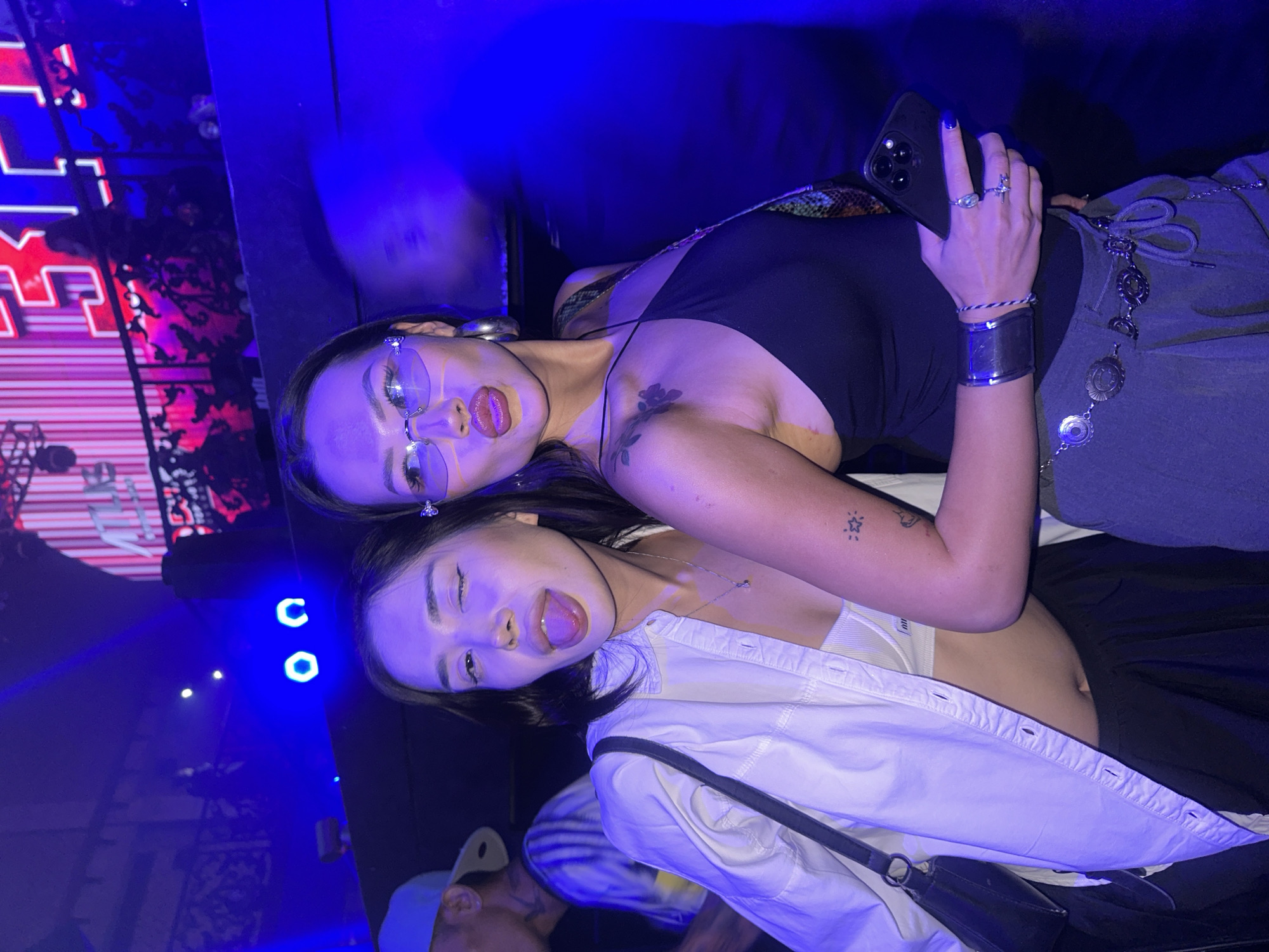 In North Kuta, Indonesia, two young women pose for a photo at a nightclub. The woman on the left, wearing a white unbuttoned shirt over a white bralette and a black skirt, playfully sticks out her tongue and winks at the camera. The woman on the right, adorned with large hoop earrings, sunglasses, and a black halter top, pouts her lips and holds a smartphone. Both women have tattoos on their arms, and the woman on the right wears elaborate silver jewelry around her waist. The background is dimly lit, with blue and pink neon lights illuminating a stage with a digital display. The atmosphere suggests a lively nightlife scene.