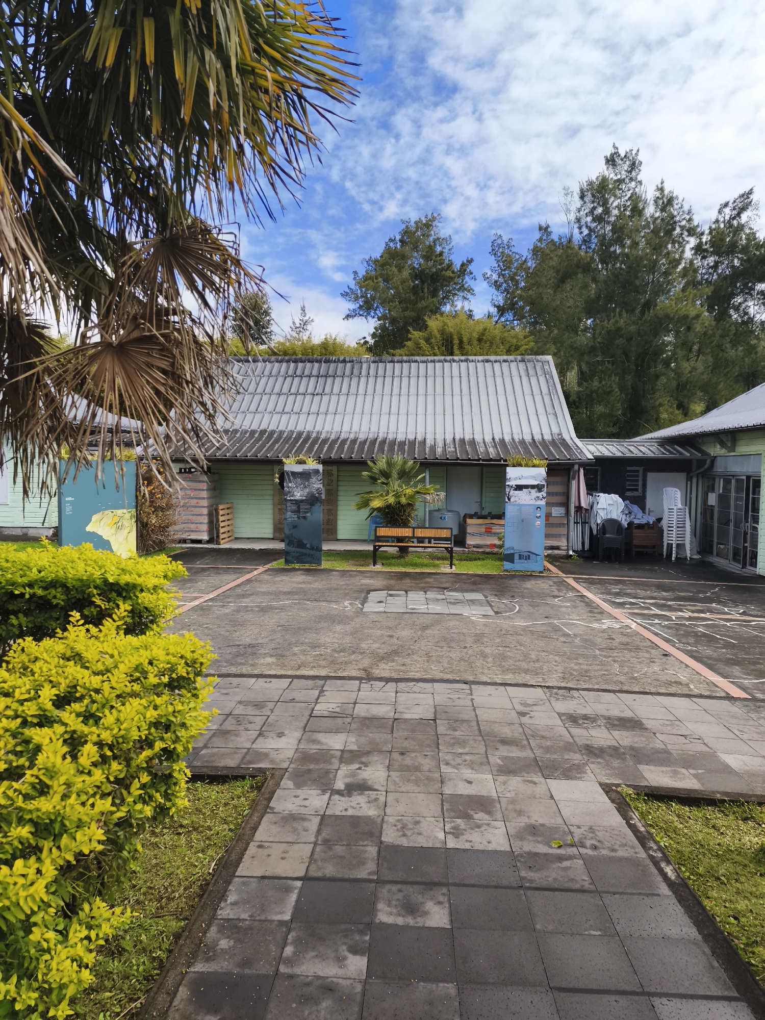 The image shows an outdoor area with a light green building and a gray, corrugated metal roof. In front of the building, there is a paved courtyard with gray square tiles. To the left, there is a vibrant yellow-green bush and a portion of a palm tree. To the right of the bush, there is a display panel with a yellow map. In the center of the paved area, there is a wooden bench. There are two tall vertical display panels in front of the building, one on either side of the bench. The building appears to be a single-story structure with multiple sections. Behind the building, there are trees and a blue sky with white clouds. There are no people visible in the image. The scene is likely in La Plaine-des-Palmistes, France, as indicated by the prompt.