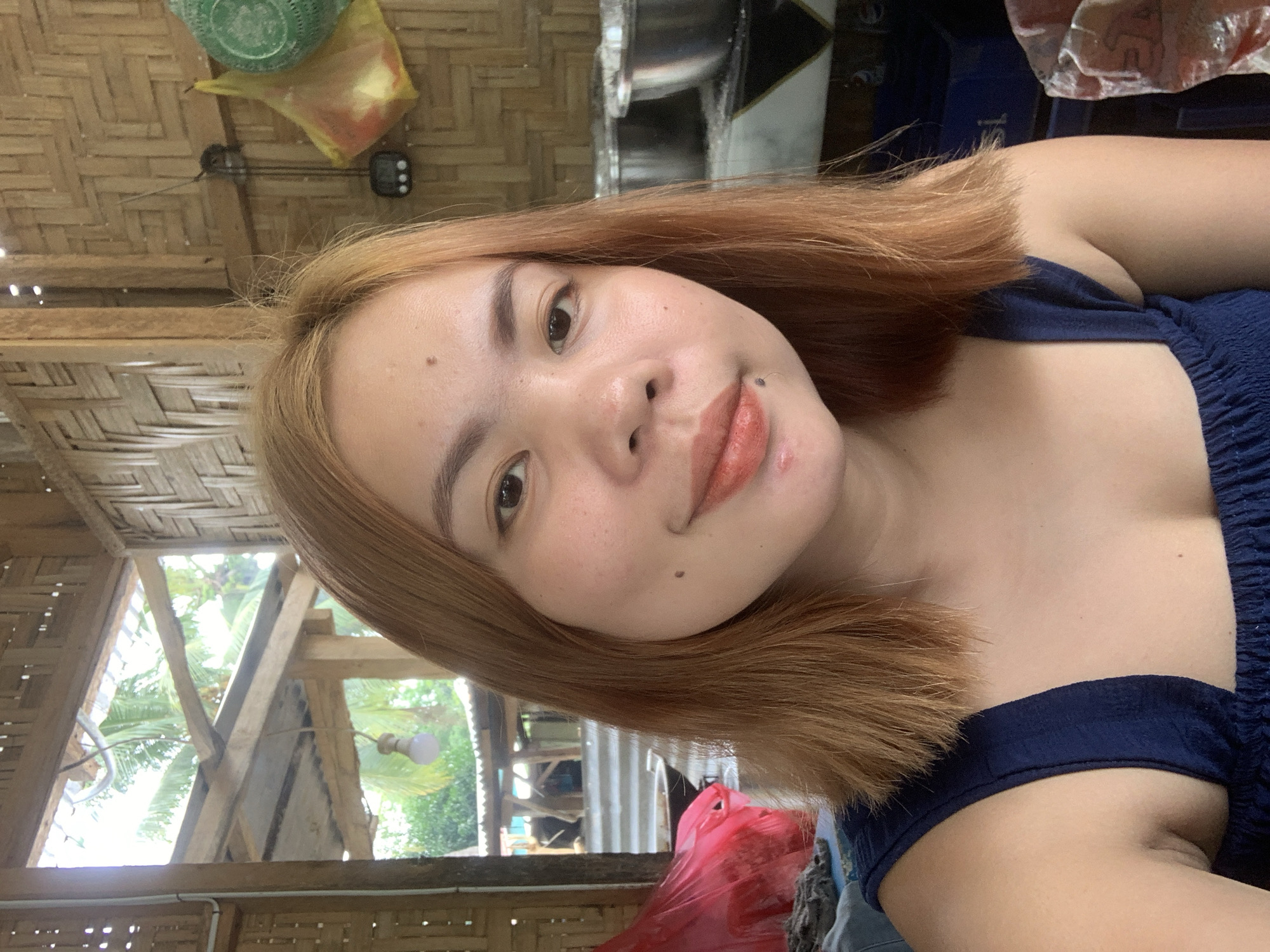 A young woman with shoulder-length auburn hair, fair skin, and subtle makeup, including lipstick and blush, looks directly at the camera. She is wearing a dark blue, ruched top. She has a small mole on her left cheek and another on her chin.
The background suggests an open-air structure with walls made of woven bamboo. Natural light filters in from an opening in the structure, revealing palm trees and foliage outside. Various objects are visible in the background, including metal bowls, a red plastic bag, and yellow and green plastic bags hanging on the wall. A light bulb hangs from the ceiling. The overall setting appears rustic and likely located in a rural or outdoor area, consistent with a location in Kiblawan, Philippines.