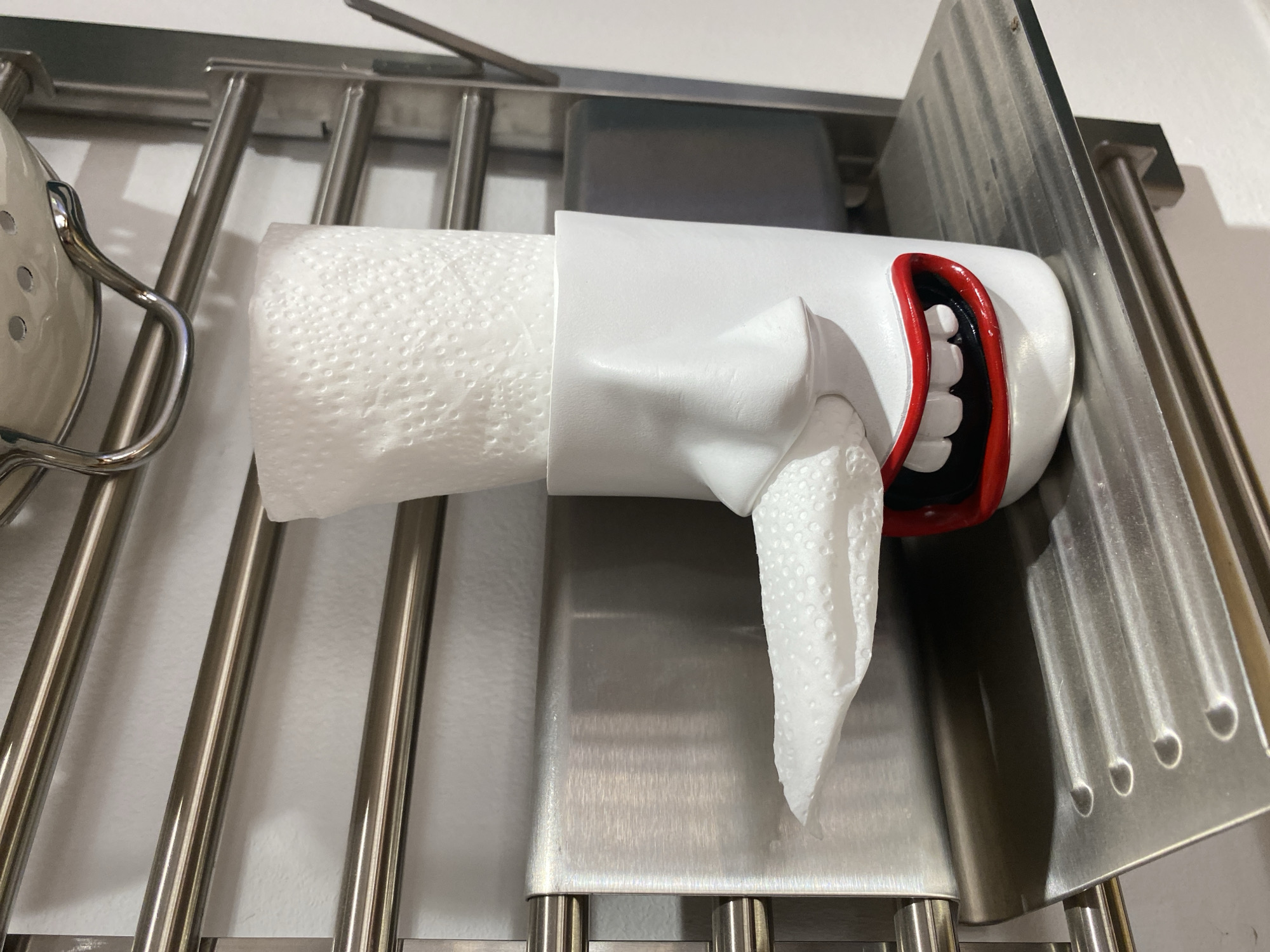 A white ceramic holder shaped like a face with a large nose and an open mouth with red lips and white teeth holds a roll of toilet paper. A piece of toilet paper is pulled out of the holder, resembling a tongue. The holder is sitting on a ribbed metal shelf. Behind the shelf is a silver towel rack with multiple bars. The wall behind the towel rack is white and textured. The overall scene is indoors, likely a bathroom or powder room. The location in Karaganda, Kazakhstan is not discernable from the image.