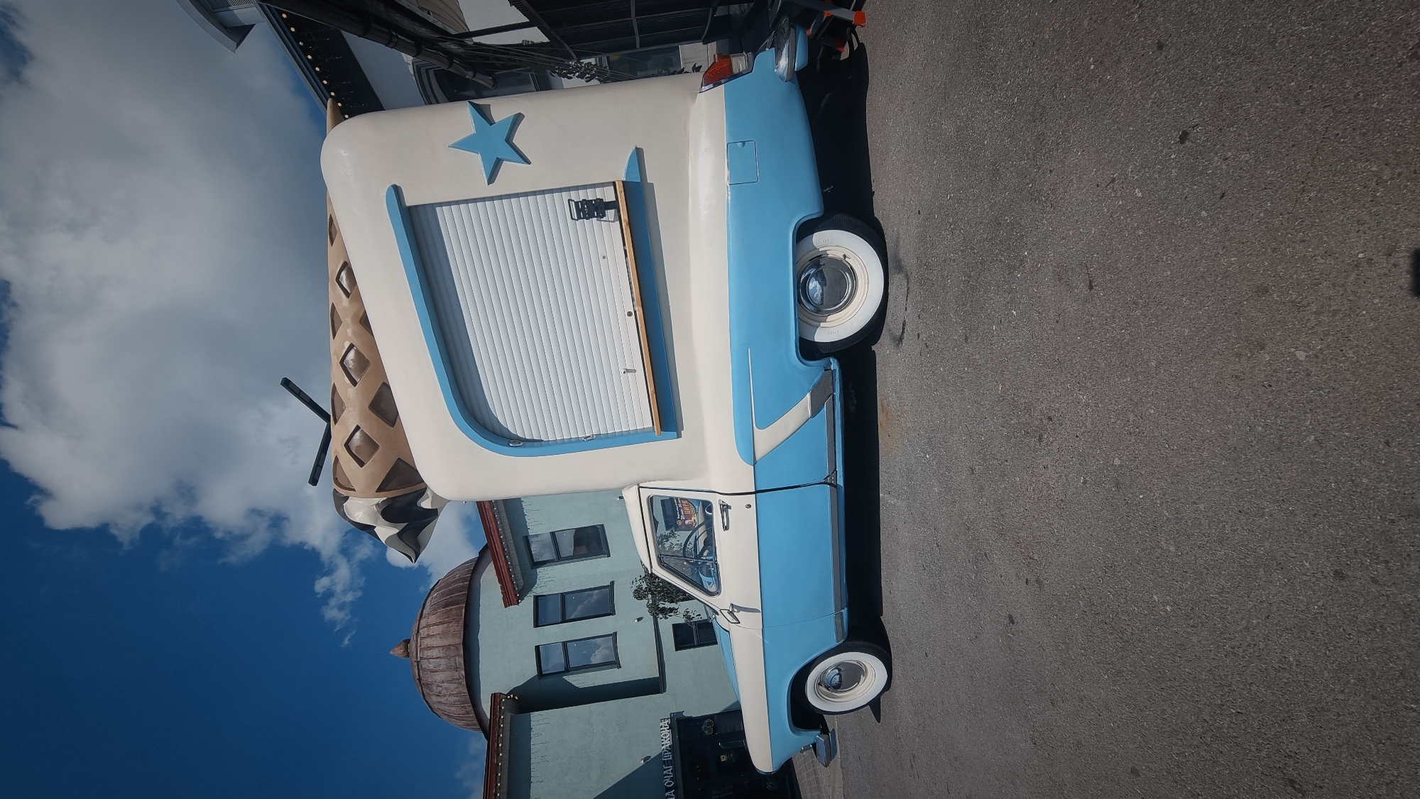An eye-level, medium shot captures a vintage blue and white pickup truck converted into a novelty ice cream truck. The truck is parked on an asphalt road in front of buildings, under a bright blue sky with scattered white clouds. The truck's rear is dramatically extended and reshaped to resemble an ice cream cone, complete with a waffle-patterned exterior and what appears to be a chocolate drizzle effect at the top, topped with black rod-like elements. A blue star emblem is affixed to the side of the extended structure, which features a closed, white roll-up door. The truck's original cab is intact, with a person visible inside, seated at the steering wheel. In the background, to the left, a light blue building with a domed roof and arched windows is visible. To the right, parts of other buildings and structures can be seen. The overall scene is vibrant and whimsical, suggesting a commercial or recreational setting. The location is identified as the city of Ufa, Russia, likely a public space or street.