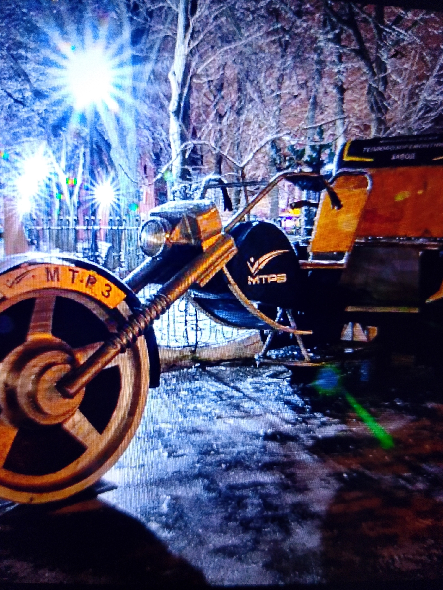 An outdoor night-time, medium shot captures a three-wheeled vehicle, possibly a motorized rickshaw, parked on a wet, possibly icy, paved surface. The vehicle has a yellow-orange rear seat section and a dark blue front body with a prominent headlight. The front wheel, partially visible on the left, is a large, spoked wheel with a yellow band and text that reads "MTR 3". A logo with stylized lines and the text "MTPS" is visible on the side of the dark blue front section. In the background, bare trees are silhouetted against bright, starburst-shaped lights. A dark fence is visible behind the trees. The ground appears wet and reflective, with hints of a green light flare towards the right side of the image. The overall scene suggests a cold, potentially urban environment, possibly a park or street in Dobra Krynytsia, Ukraine.