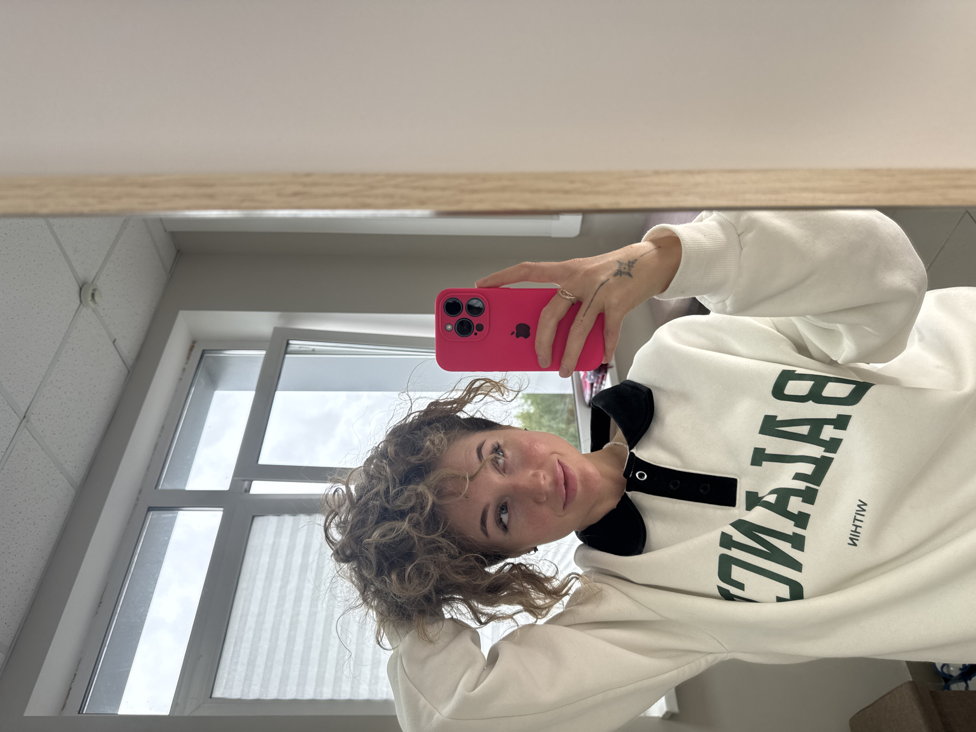 A selfie in a mirror shows a woman with curly, light brown hair. She is holding a pink iPhone in her right hand, adorned with a ring and a tattoo of a star-like symbol on her wrist. She is wearing a white sweatshirt with a black collar and a green inscription that reads "BALENCIAGA" reflected in the mirror. The background reveals a room with a window. Outside the window, the sky appears cloudy. A fluorescent light is visible on the ceiling. The location in the city of Chaikovsky, Russia cannot be determined from the image alone.