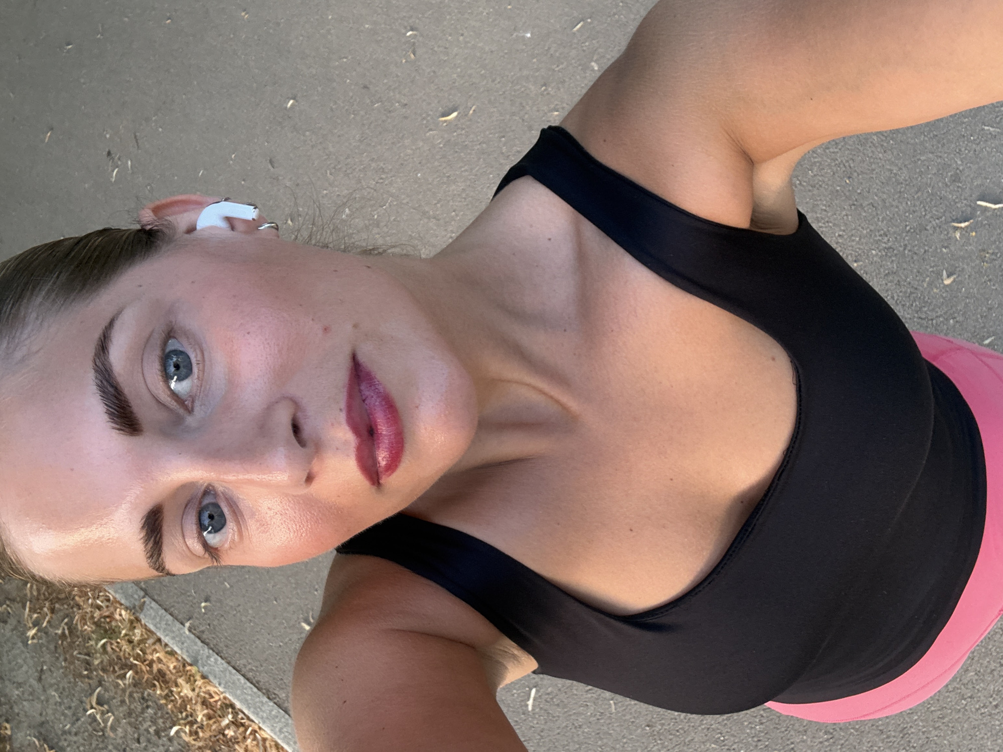 The image shows a close-up, high-angle shot of a woman outdoors. She has fair skin, blue eyes, and dark eyebrows, and is wearing dark lipstick and a black tank top. A white earbud is visible in her right ear, and a silver hoop earring is in her left earlobe. Her hair is pulled back. The woman is standing on a paved surface, possibly a sidewalk or path, with some dry leaves and debris visible on the ground. To the left of the path, there is an area covered in dry vegetation and dirt. The lighting suggests it is daytime, possibly late afternoon or early morning, given the shadows and the quality of light on her skin. There is no indication of the location being in Paris, France.