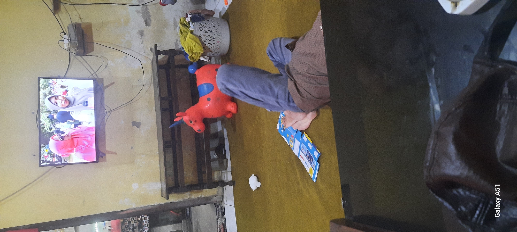 In the city of Jatitengah, Indonesia, a dimly lit, somewhat cluttered room is depicted. A large, flat-screen television mounted on the wall displays an image of two women wearing headscarves. Below the television, a long, wooden cabinet with carved legs stands on the floor. To the right of the cabinet, an orange inflatable horse toy with a blue saddle is present. A white laundry basket filled with clothes sits next to the toy. In the foreground, a person's legs and feet are visible, with the person lying on a brown rug. A colorful booklet lies open on the rug near the person's feet. A small, white object, possibly a toy, is also on the rug. The walls of the room are discolored and appear to be peeling in places. Electrical wires are visible, connected to a power strip on the wall. The scene suggests a casual, domestic setting.