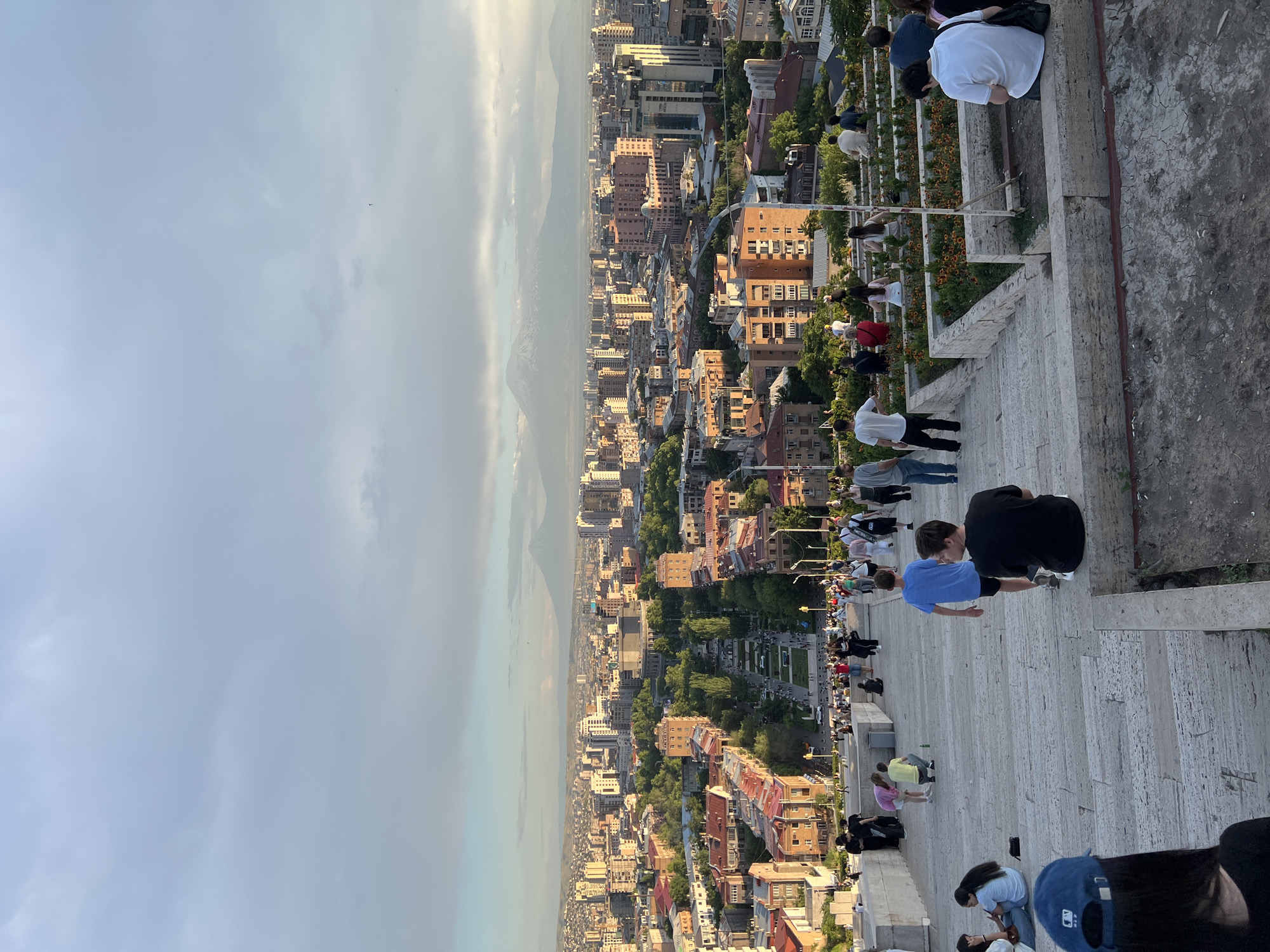 The image depicts a panoramic view of Yerevan, Armenia, from an elevated vantage point, likely the Cascade Monument, overlooking the city and the distant Mount Ararat. In the foreground, a wide, stone staircase is populated by numerous individuals in casual attire, some seated, others standing or walking. The stairs are constructed from large, light-colored stone blocks.
Further down the staircase, a park-like area with flowerbeds is visible, also with people gathered. Beyond this, the cityscape unfolds, characterized by a dense arrangement of buildings with terracotta roofs and a variety of architectural styles, interspersed with lush green trees. Prominent structures, possibly cultural institutions or large apartment complexes, rise above the general urban fabric.
In the far distance, the majestic silhouette of Mount Ararat, capped with snow, is discernible against a hazy, cloudy sky, suggesting it is either early morning or late afternoon. The sky itself is a blend of pale blues and grays, with soft light filtering through the clouds, creating a serene atmosphere. The overall scene captures a moment of urban life and natural beauty in Yerevan.