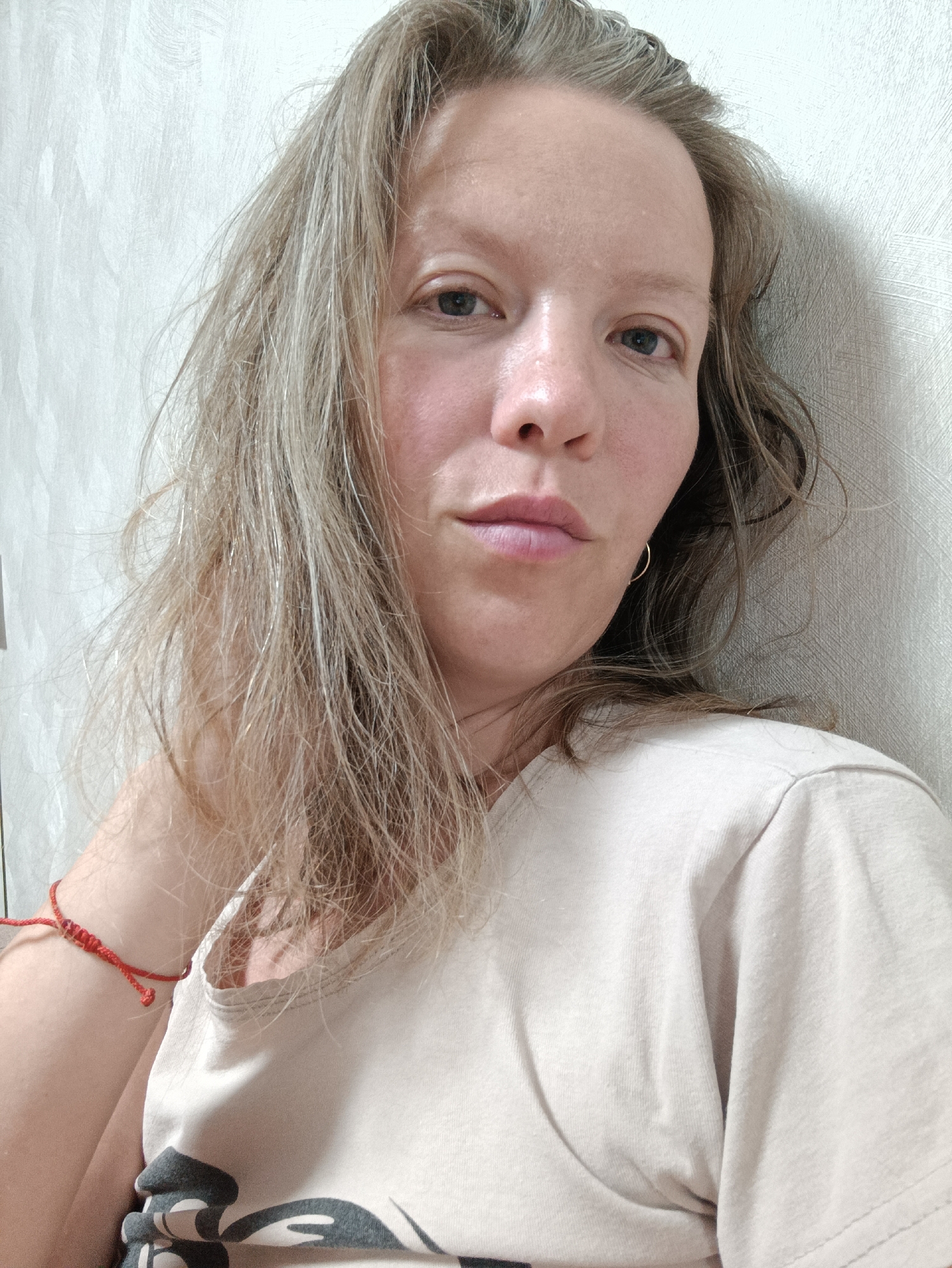 A woman with light brown, shoulder-length hair is close to a textured, off-white wall, likely indoors. She is wearing a light beige t-shirt, and a thin red bracelet is visible on her left wrist. A small gold hoop earring is on her right ear. The woman's complexion is fair. The picture's location is Chaikovsky, Russia.
