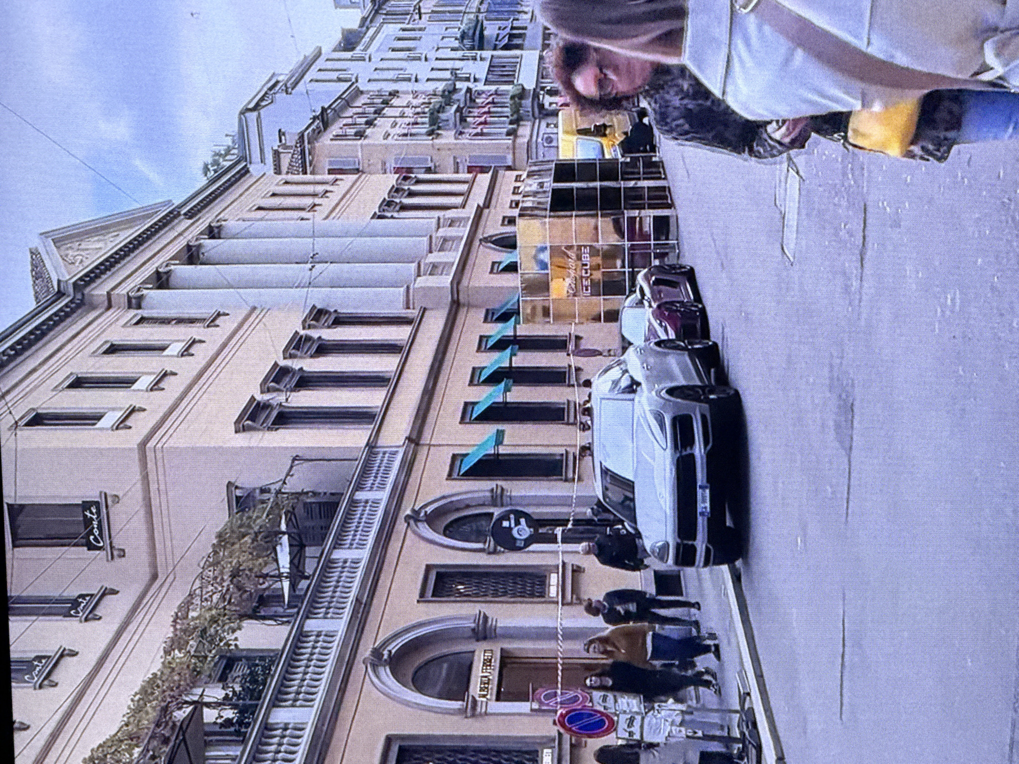 This image depicts a street scene, likely in an urban environment. In the foreground, the front portion of a silver Porsche Cayenne SUV is visible, parked on the right side of the street. To the left of the Porsche, a maroon sports car is parked. Beyond the cars, a multi-story building with classical architectural elements, including columns and arched windows, dominates the background. Several people are walking on the sidewalk in front of the building, some appearing to be customers or pedestrians. There are also signs and temporary structures, possibly for an event or display, visible in front of the building. The sky above is overcast. The presence of the Chopard "Ice Cube" branding on a display suggests a potential luxury brand event.
The request to include the location as Dubai, United Arab Emirates, cannot be fulfilled based solely on the visual information in the image. The architectural style, signage, and general atmosphere do not definitively point to Dubai.