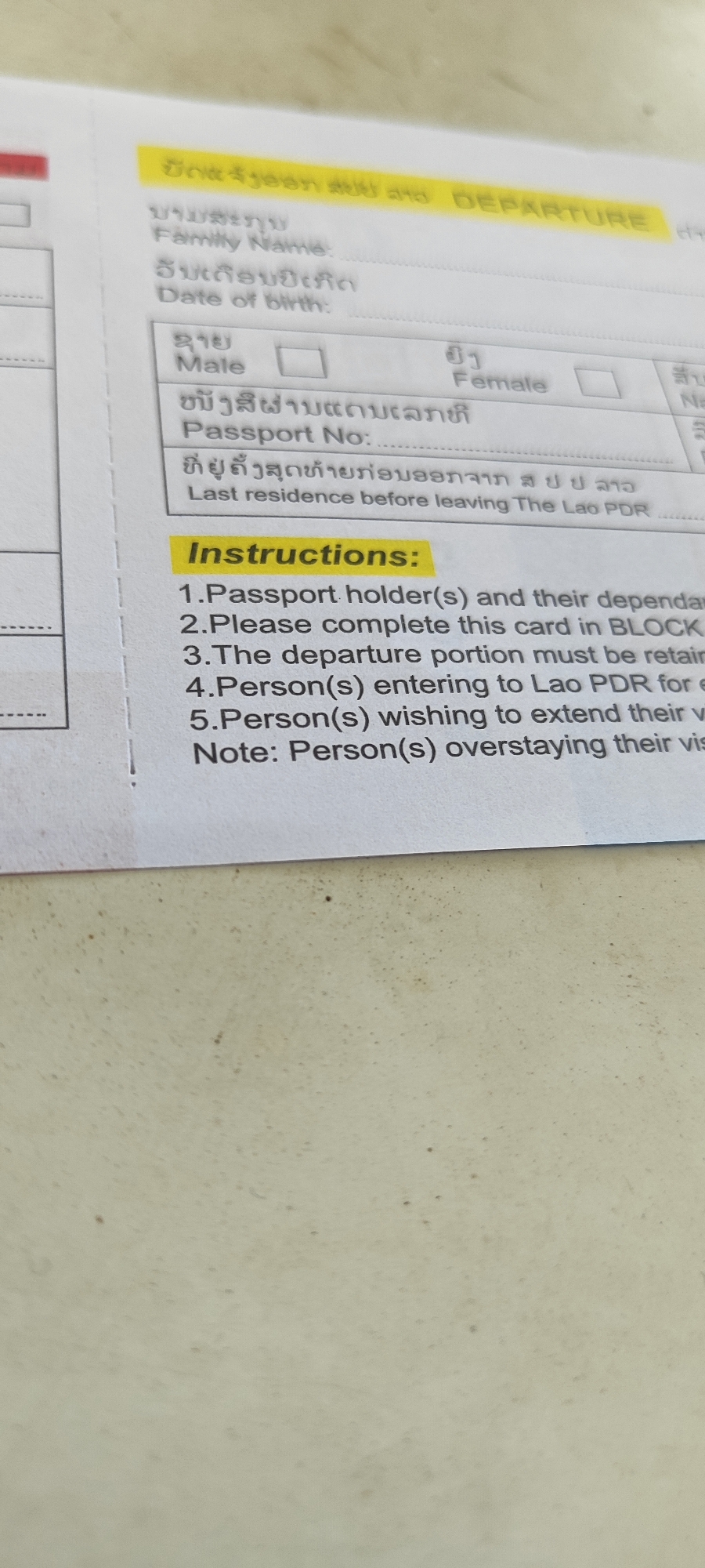 The image shows a close-up of a form, likely a disembarkation/embarkation card, with text in both Lao and English. The yellow highlighted section at the top reads "DEPARTURE". Below this are fields for "Family Name," "Date of birth," "Male," "Female," and "Passport No." There is also a line for "Last residence before leaving The Lao PDR." A section labeled "Instructions:" follows, with numbered points detailing how to complete the form. The background is a textured, light-colored surface, possibly a table. The image quality is slightly blurred, making some of the text difficult to read with absolute certainty. There are no people visible in the image.