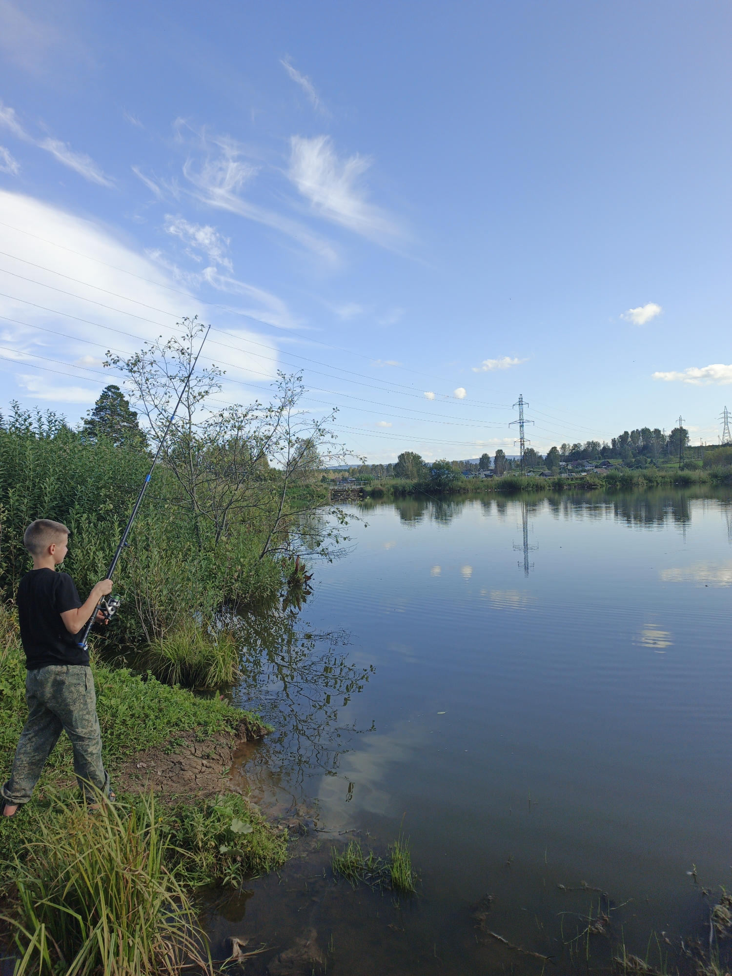 A young boy is fishing on the bank of a calm body of water in Urtiushka, Russia. He is wearing a black t-shirt and camouflage pants, and is holding a fishing rod. The water is reflecting the blue sky and white clouds, as well as the surrounding trees and power lines. The vegetation on the bank is lush and green, with some dry, cracked earth visible. In the distance, there are power lines and a line of trees on the horizon. The sky is a bright blue with wispy white clouds.