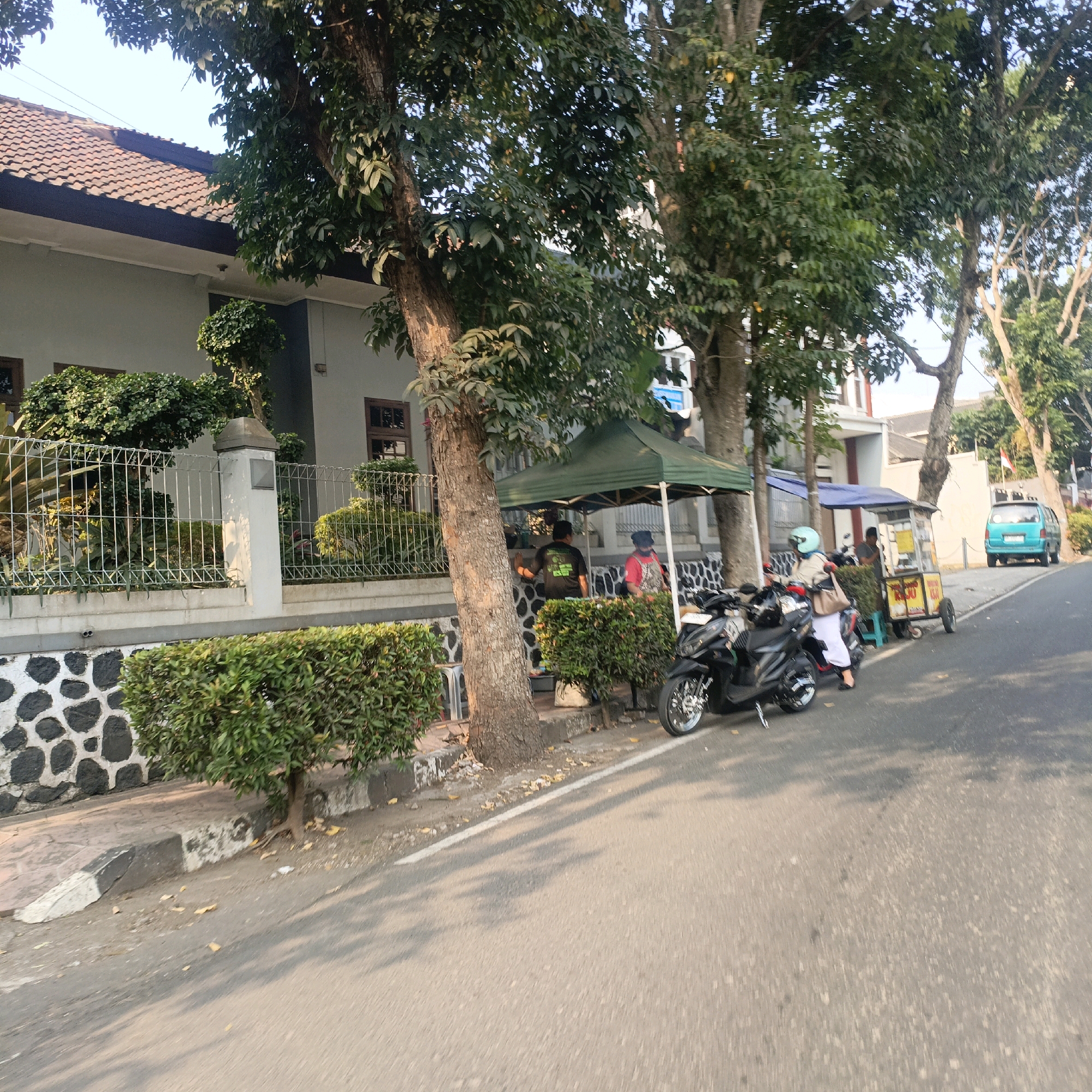 The image depicts a street scene in Salatiga, Indonesia, featuring a residential building with a white facade and a tiled roof. In front of the building is a meticulously maintained garden with hedges and ornamental plants, enclosed by a metal fence and a stone wall. Several trees with lush green foliage line the street, casting shadows on the pavement.
A green pop-up tent is set up on the sidewalk, serving as a temporary stall. Two individuals are visible near the tent; one, wearing a black t-shirt with green lettering, appears to be interacting with someone inside the tent, while another person, in a red shirt and a head covering, is also near the stall.
Further down the street, another stall is visible, this one a mobile cart with yellow and red signage, and a blue awning. A person wearing a light blue helmet and a white outfit is seated on a black motorcycle parked next to the cart. Another black motorcycle is parked in front of the first stall.
A bright teal colored van is parked on the right side of the road, further down the street. The road itself is paved and appears to be a typical urban street. The overall scene suggests a lively street-side activity, possibly related to local vendors or small businesses operating along the residential area.
