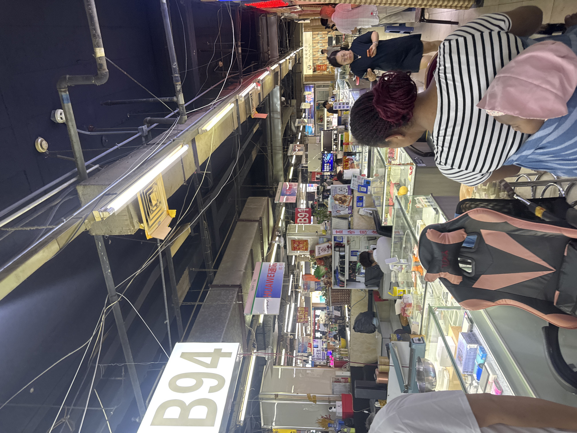 The image depicts an indoor marketplace, likely an electronics market, in Dadong District, China. A large sign with "B94" is prominently displayed overhead. The ceiling is a dark expanse with numerous exposed electrical wires and conduits, along with fluorescent lighting fixtures. Various stalls are visible, showcasing goods behind glass display cases. One stall has a sign that says "HUAWEI 通讯" (HUAWEI Communications). In the foreground, a woman with braided hair is carrying a baby in a sling. She is wearing a striped shirt. To her right, another woman in a black dress is holding a smartphone. A man in a plaid shirt stands to the right of the woman in the black dress, facing away from the camera. A gaming chair with pink accents is positioned in the lower left of the frame. The overall environment suggests a busy commercial setting with a variety of products and people engaged in shopping or work.