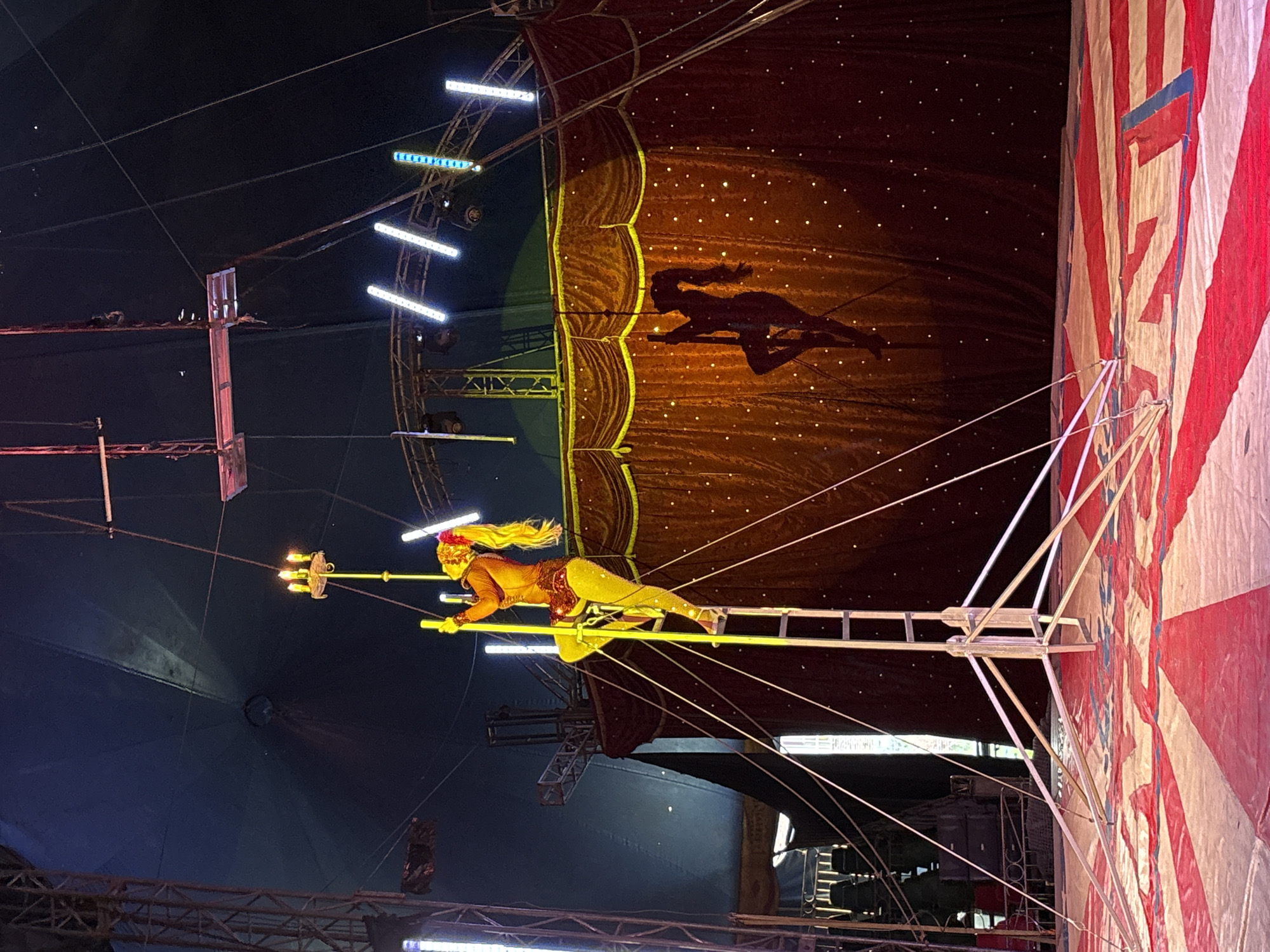 The image depicts a circus performance in Tejalpa, Mexico. A female performer, dressed in a yellow and gold costume with a feathered headdress, balances on a high-wire ladder contraption. She holds a long pole with lit torches at the end. The performance takes place inside a traditional red and white striped circus tent. The background includes rigging, lights, and a decorative curtain with a silhouette of a horse and rider.