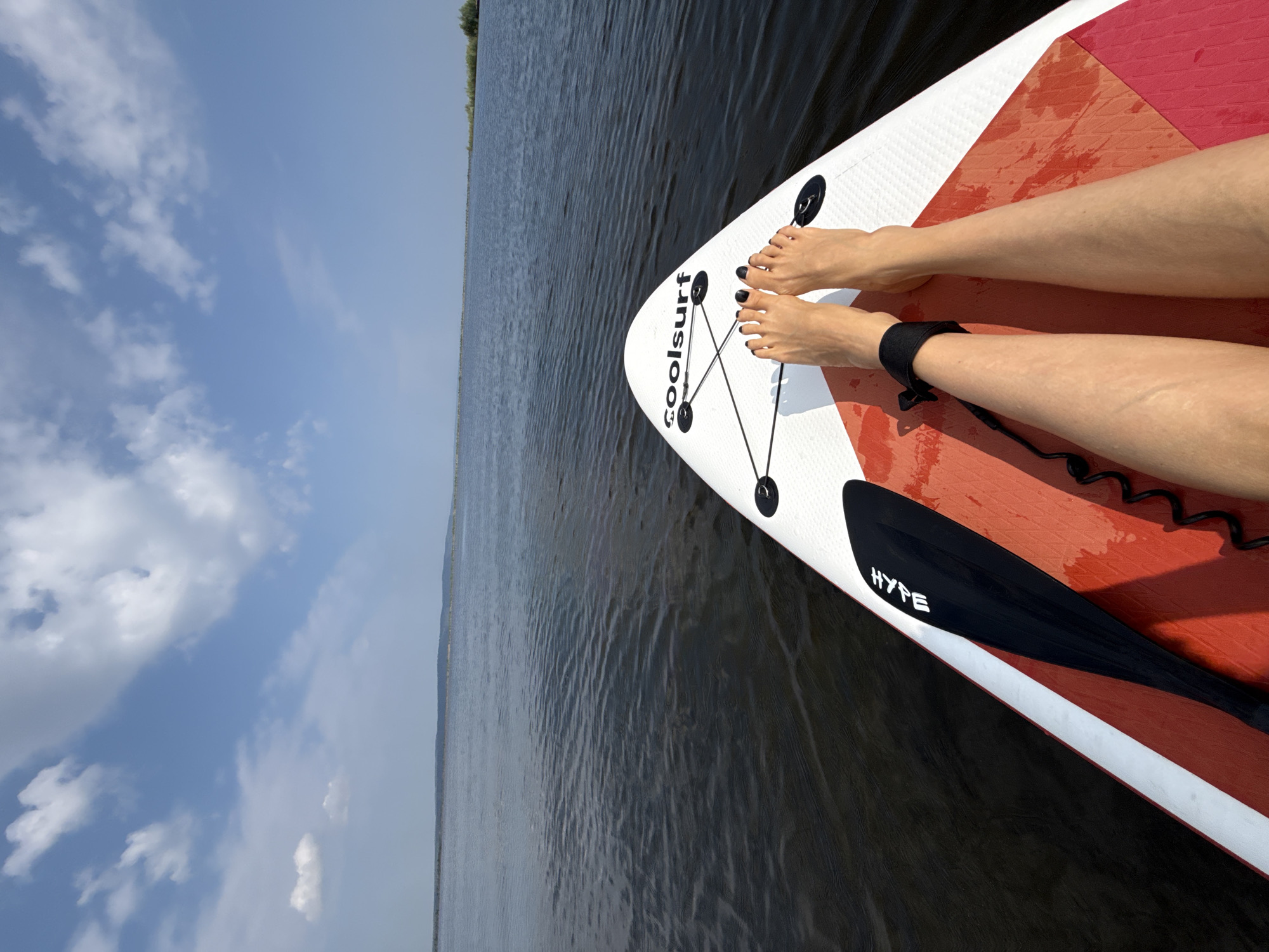 A first-person perspective shot captures a person's legs and feet resting on a white and red paddleboard on a calm, dark body of water. The paddleboard, which has the brand name "Coolsurf" printed on it, is equipped with a black paddle with "HYPE" written on it, and a black ankle leash is attached to the person's leg. The water is still, with subtle ripples reflecting the bright blue sky filled with scattered white clouds. In the distance, a hazy, tree-lined horizon stretches across the frame. The image appears to be taken from the shore of the Kama River or a similar large water body near Chaikovsky, Russia, given the expansive and serene nature of the scenery.