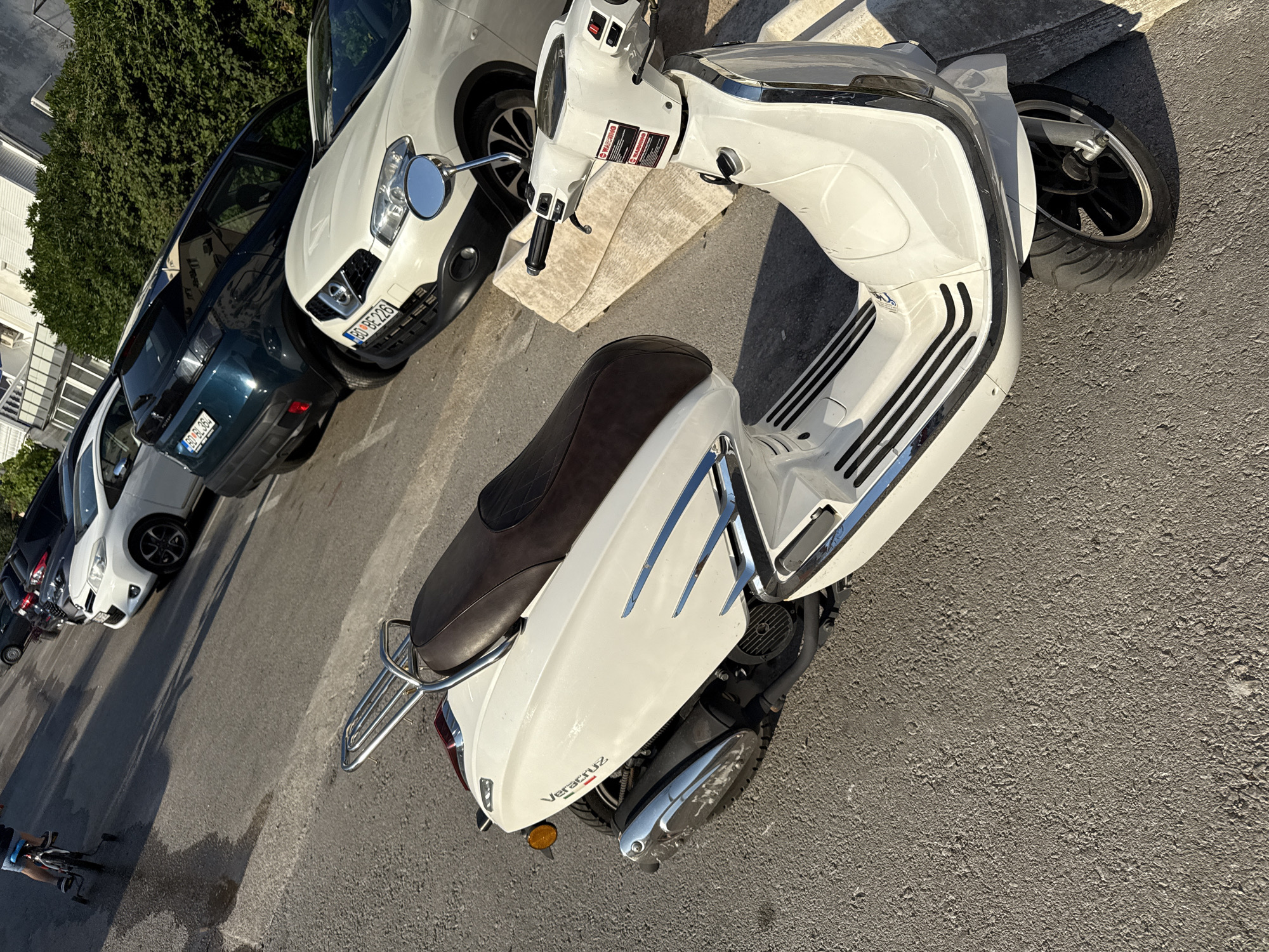 A white scooter is parked on an asphalt surface, angled towards the right of the frame. The scooter has a brown seat and chrome accents. To the left of the scooter, a person on a bicycle with training wheels rides on a wet patch of asphalt. In the background, several cars are parked along the street, including a dark green Peugeot and a white Nissan. Buildings with white facades and dark windows are visible behind the parked cars. The scene appears to be a street in Budva, Montenegro, with buildings and parked vehicles suggesting an urban environment. The lighting indicates it is daytime, with shadows cast across the asphalt.