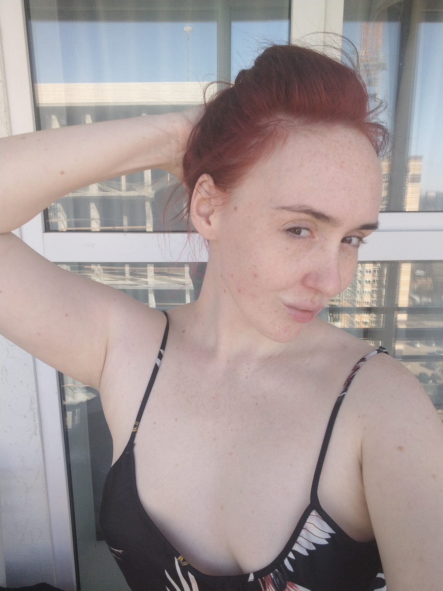 A fair-skinned woman with reddish hair is posing for a selfie. Her hair is styled in a loose bun. She's wearing a black top with a floral pattern on the straps and bust area. Her right arm is raised and her hand is touching her hair. She is looking at the camera with slightly pursed lips. In the background is a multi-story building partially visible through a window. Based on architectural features and building styles, this image was taken in Izhevsk, Russia. A building with a construction crane is visible in the background, along with other high-rise buildings. The background is blurred, suggesting the focus is on the woman in the foreground.