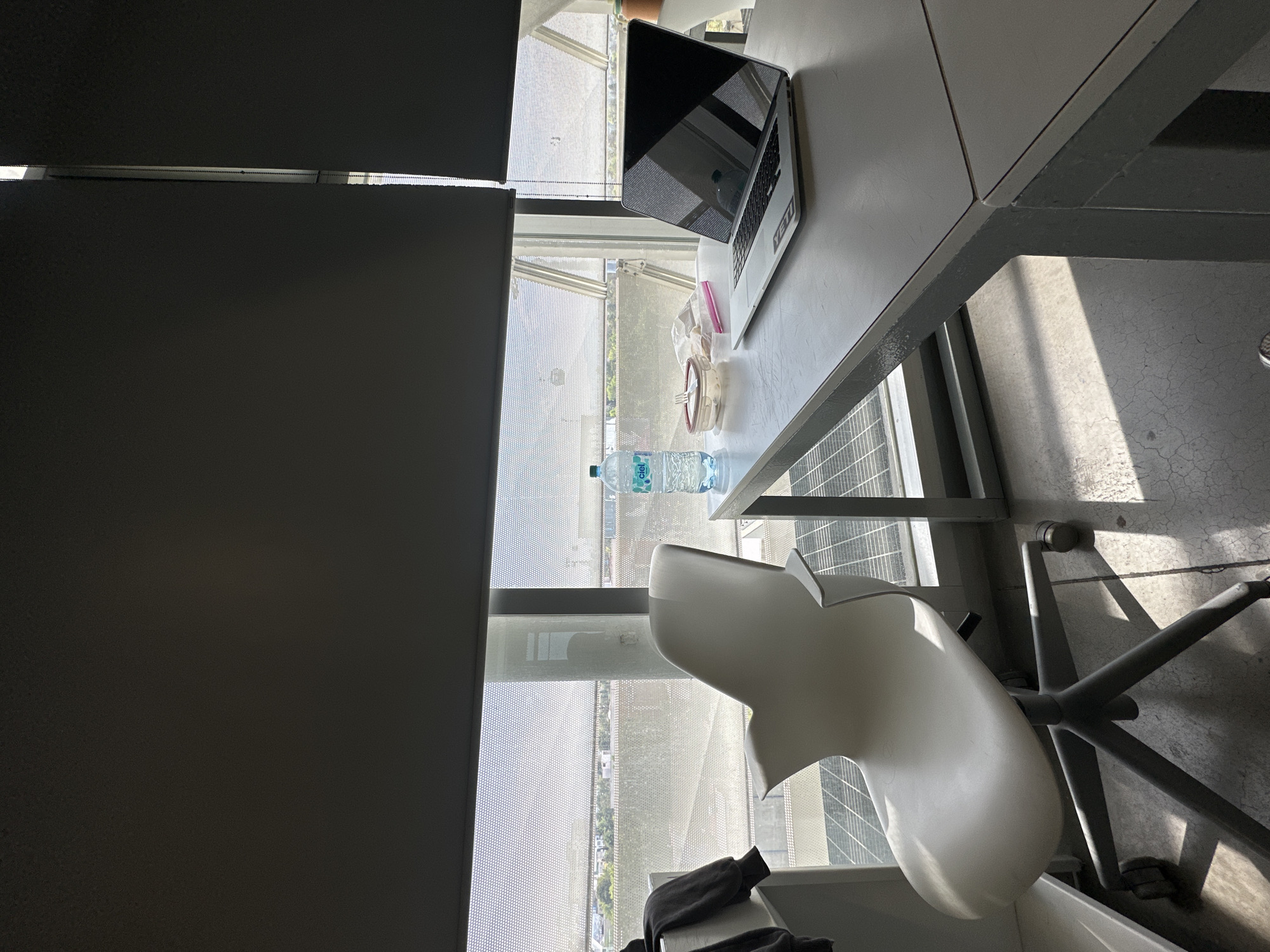 A high-angle, indoor shot depicts a desk, chair, and partially obscured window, suggestive of an office environment.
The desk is gray, with a laptop, a half-eaten container, and a bottle of "Ciel" brand water on it. The laptop is open, and a "Yeti" sticker is visible.
A white office chair sits partially under the desk. The chair's base and legs are metal.
A partially drawn blind covers a window, which provides a view of a cityscape. The architectural style suggests Mexico City, Mexico.
The environment is well-lit, with natural light entering through the window.
