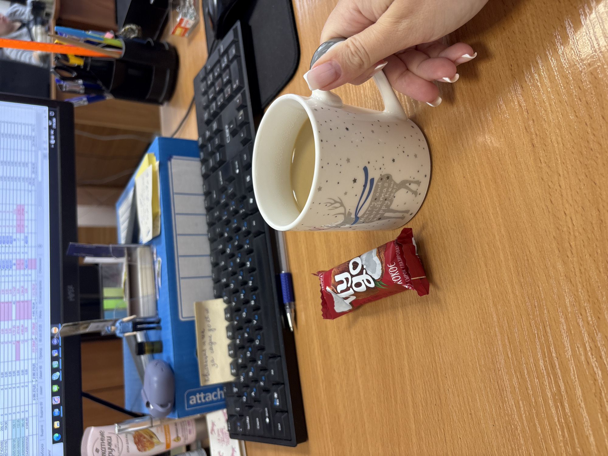 Here is a detailed description of the image:
**Environment:**
* The scene appears to be set on an office desk, with a wood-patterned surface.
**Subjects & Objects:**
* A human hand with light skin and French manicure nails is holding a white ceramic mug with a design featuring reindeer, snowflakes, and the text "Merry and Christmas happy new year". The mug contains a light-colored liquid, likely coffee or tea.
* A black computer keyboard is visible, positioned horizontally on the desk.
* A computer monitor displaying what appears to be spreadsheet data is positioned on the left side.
* A blue office organizer is next to the keyboard and monitor
* A blue ballpoint pen lies near the keyboard and a package of coconut candy
* Several items are situated in the background, including office supplies (pens, paper clips), hand cream and box of cookies
* A candy bar, labeled "Кокос" (Coconut), lies on the desk near the mug.
**People:**
* There are no full human figures visible, but a hand is holding the mug.
**Actions:**
* The hand is holding the mug, presumably for drinking.
**Setting:**
* The scene could be set in a commercial building in Rusanovo, Russia due to the candy bar's Russian labeling.
**Note:** The exact location is impossible to verify beyond the probability of being in Russia, due to the Russian text on the candy bar.