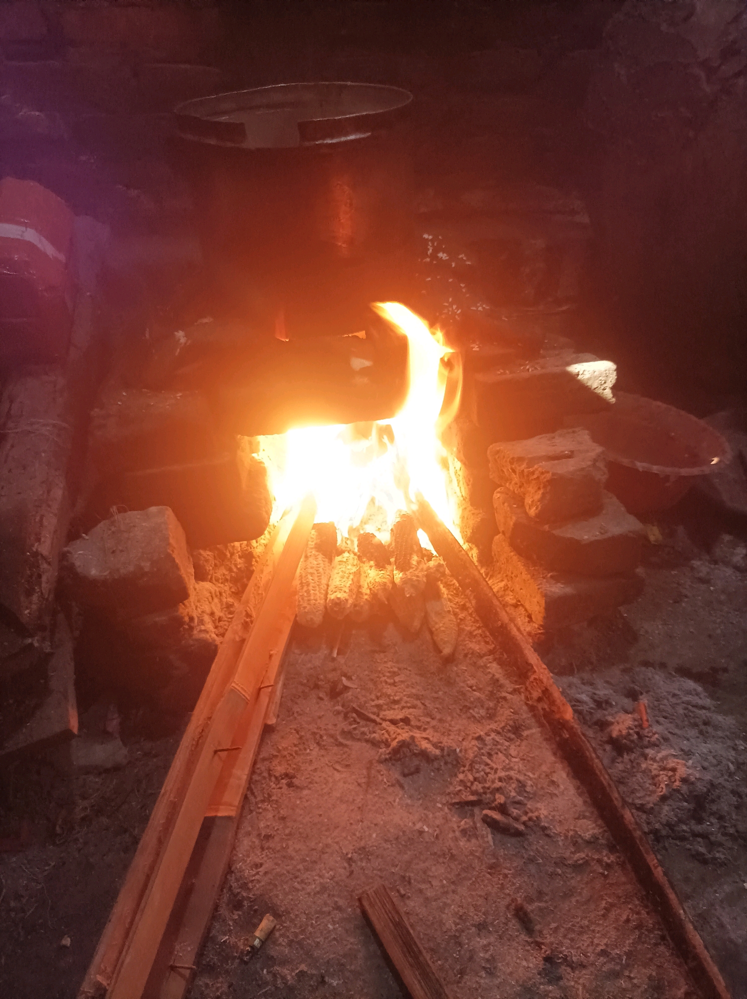 In Canga'an, Indonesia, a roaring fire burns brightly in a rustic outdoor cooking area. The fire is contained within a makeshift structure of bricks and rough-hewn wood. Several ears of corn are being roasted directly over the flames, arranged on wooden supports. Above the fire, a large metal pot or cauldron is suspended, hinting at food preparation on a larger scale. The surrounding environment is earthy and somewhat unkempt, with ash and debris scattered around the cooking area. The lighting is dominated by the warm, flickering glow of the fire, casting deep shadows and highlighting the textures of the materials.