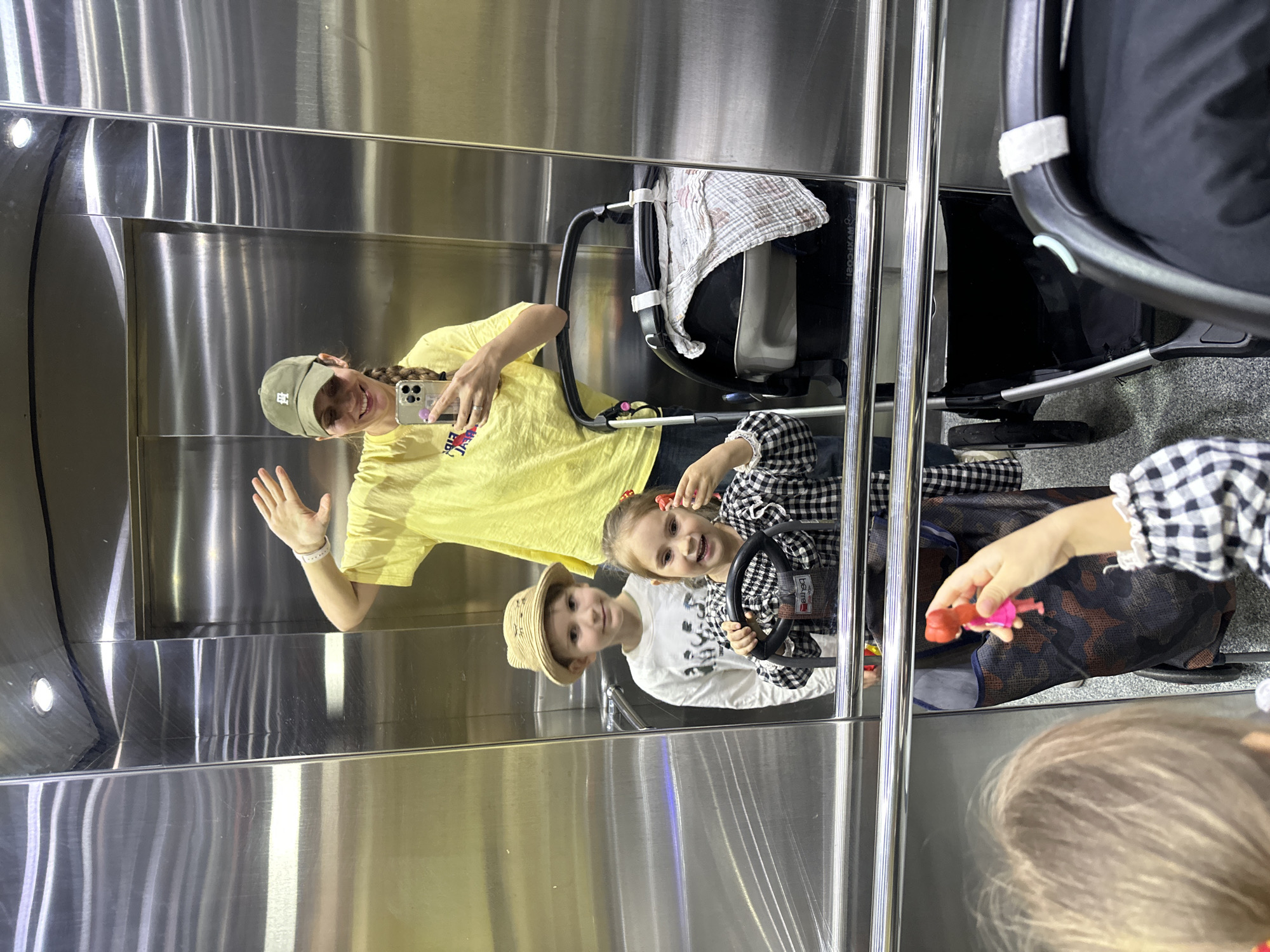 A woman with long braided hair wearing a yellow T-shirt and a baseball cap is taking a selfie in a stainless steel elevator. She is waving with her left hand. Two young children are also visible in the elevator. One child is wearing a white shirt and a tan hat, and the other is wearing a black and white checkered shirt and has red bows in her hair. The checkered-shirted child is holding a small pink doll in her right hand and has her left hand on a toy steering wheel. A baby stroller is also in the elevator. The reflection of the people is visible on the elevator doors. The location is likely Fethiye, Turkey.