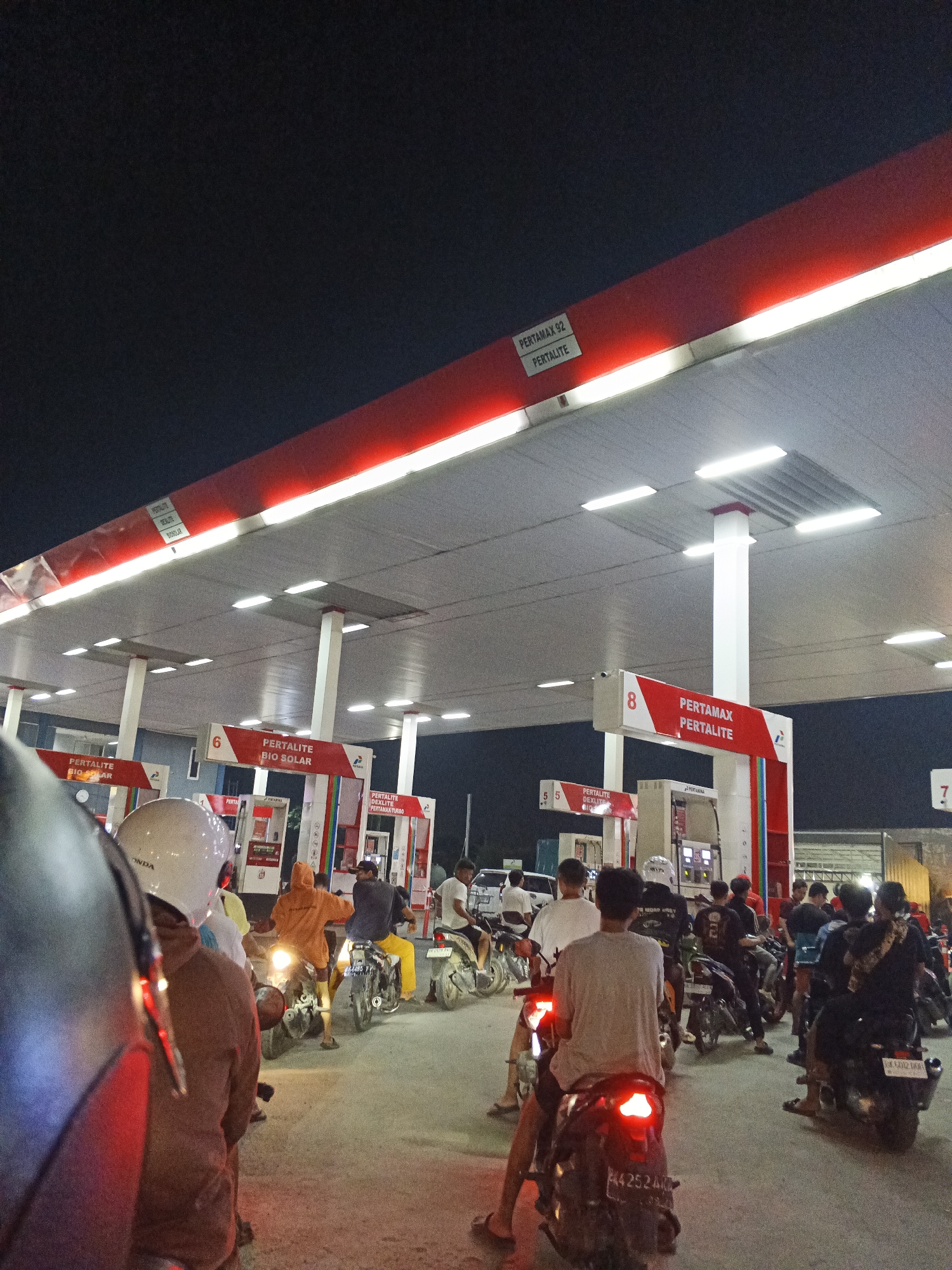 The image captures a nighttime scene at a Pertamina gas station in Ujung Serdang, Indonesia. Several motorcycles and a few cars are present, with people queuing to refuel. The gas station canopy is illuminated by bright fluorescent lights and features red and white signage indicating fuel types such as "Pertamax 92," "Pertalite," and "Bio Solar." The atmosphere appears busy, with individuals on their motorcycles waiting their turn. Some are actively refueling, while others are parked or moving through the station. The road surface is paved, and the surrounding environment is dark, characteristic of nighttime in a potentially urban or suburban setting.