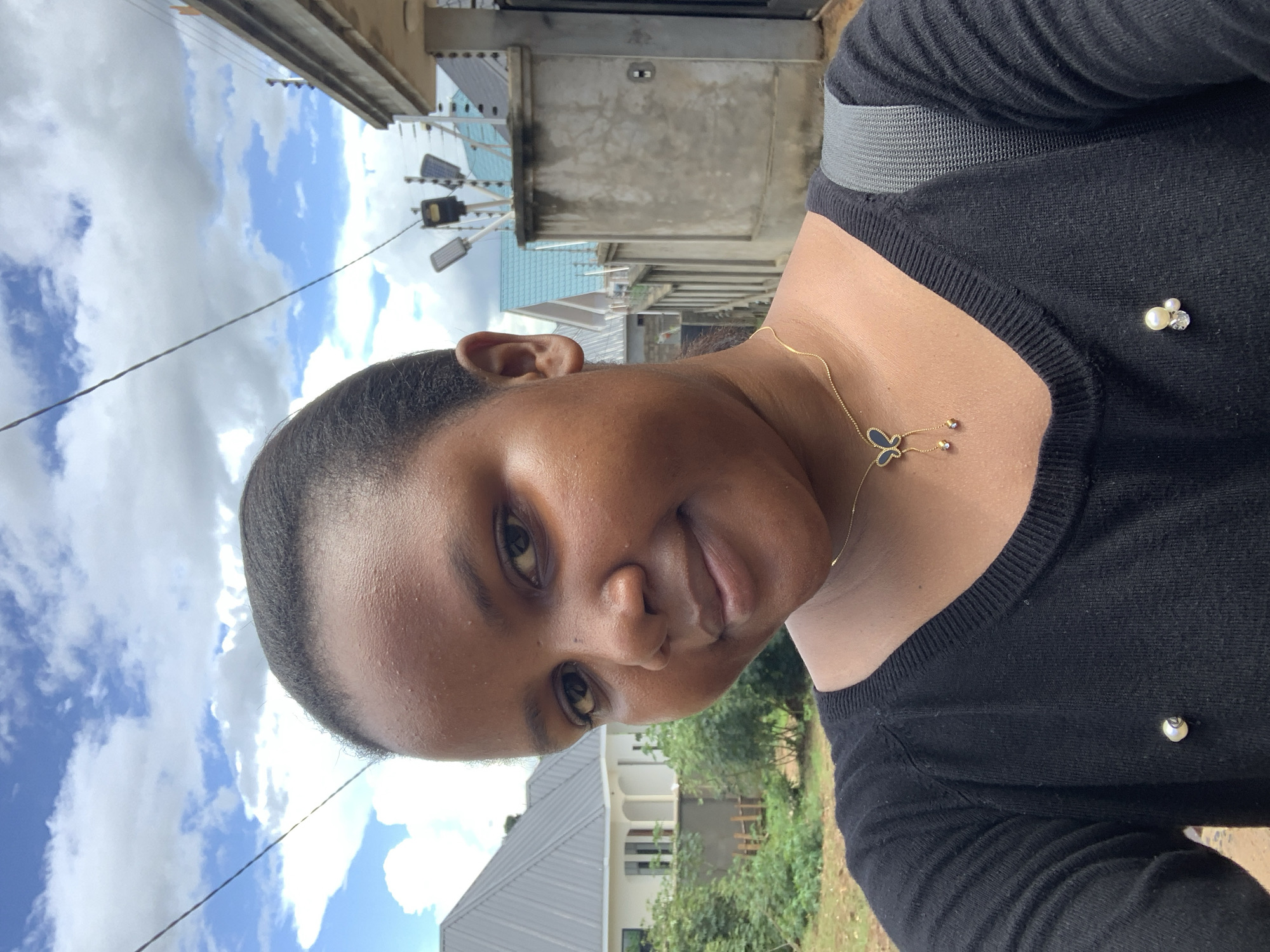 The image displays a close-up, selfie-style portrait of a female individual with a dark complexion, centered, looking directly at the camera with a slight smile. She is wearing a black long-sleeved top. A gold-colored necklace with a dark butterfly-shaped pendant, featuring two dangling elements with small beads or crystals, is visible around her neck. Two decorative pins are affixed to her upper left chest: one is a single pearl, and the other is a cluster of pearls and clear faceted stones. A grey backpack strap is visible on her right shoulder.
The outdoor background features residential buildings and vegetation under a partially cloudy sky with visible blue patches. To the subject's left, a modern, light-colored residential building with a grey corrugated metal roof and white walls is visible behind green foliage and shrubs. A small, low wooden fence is partially obscured by the vegetation near the base of this building. To the subject's right, a concrete perimeter wall topped with electric fence insulators and wires runs horizontally. Behind this wall, multiple residential structures with blue and grey sloped roofs are visible, along with several pole-mounted streetlights, which appear to be solar-powered. The architectural styles, presence of security fencing, and type of vegetation are consistent with residential areas in Dar es Salaam, Tanzania, indicating a suburban or peri-urban environment within the city.
