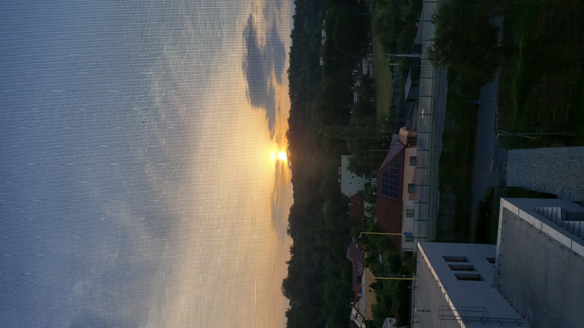 The image shows a sunset viewed through a window screen. In the foreground, there are parts of two buildings, one is a light gray, modern structure with rows of windows, and another is a lighter gray flat-roofed structure with diagonal lines. In the middle ground, there is a residential area with several houses. One house in particular is prominent, with a red roof and solar panels. Beyond the houses, a dark, tree-covered hillside forms the background, with the setting sun positioned just above the crest of the hill, casting a warm orange glow. A few faint clouds are visible in the sky. The scene is one of suburban tranquility at dusk. The approximate location in Prešov, Slovakia, can be inferred from the architectural style of the buildings and the surrounding landscape, suggesting a view from a building overlooking a residential area on the outskirts of the city.