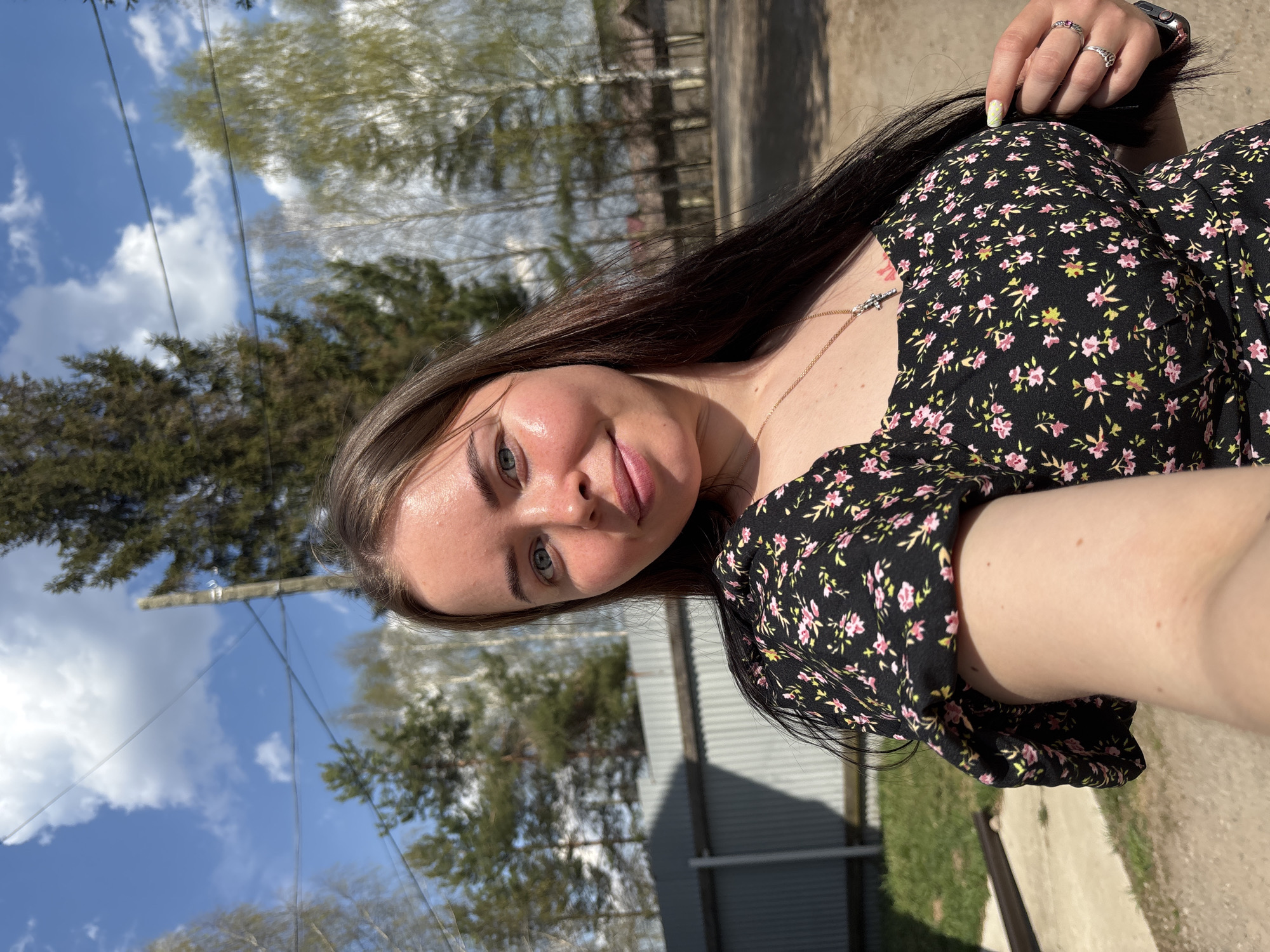 A young woman is posing for a selfie outdoors. She is wearing a dark, floral-patterned dress and has long brown hair. A silver necklace with a cross pendant is visible around her neck, and she is wearing multiple rings on her fingers. In the background, there are trees and a building with a corrugated metal exterior. The sky is blue with scattered clouds. The location may be the city of Русский Вожой, Russia.
